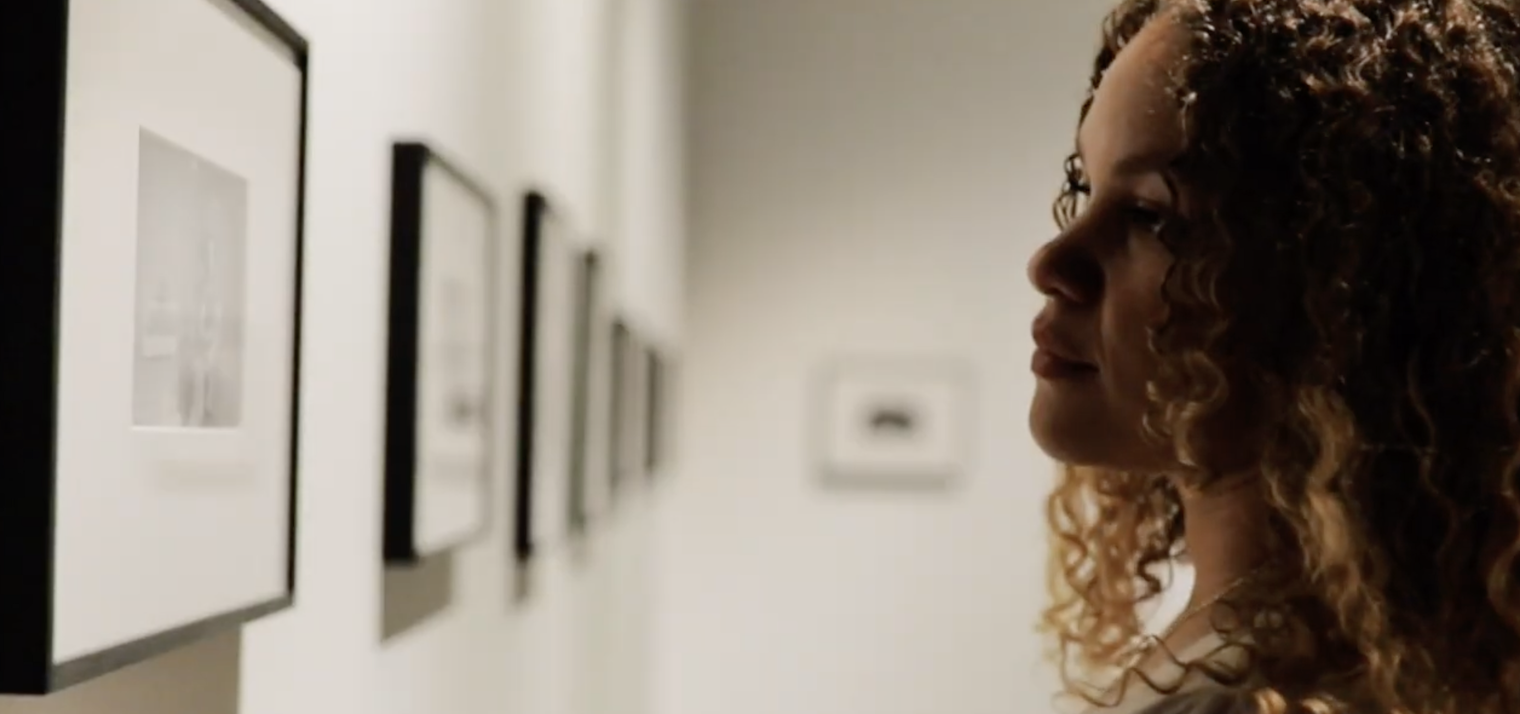Een vrouw met gekrulde, bruinrode haar kijkt naar opgehangen kunstwerken in een galerij.