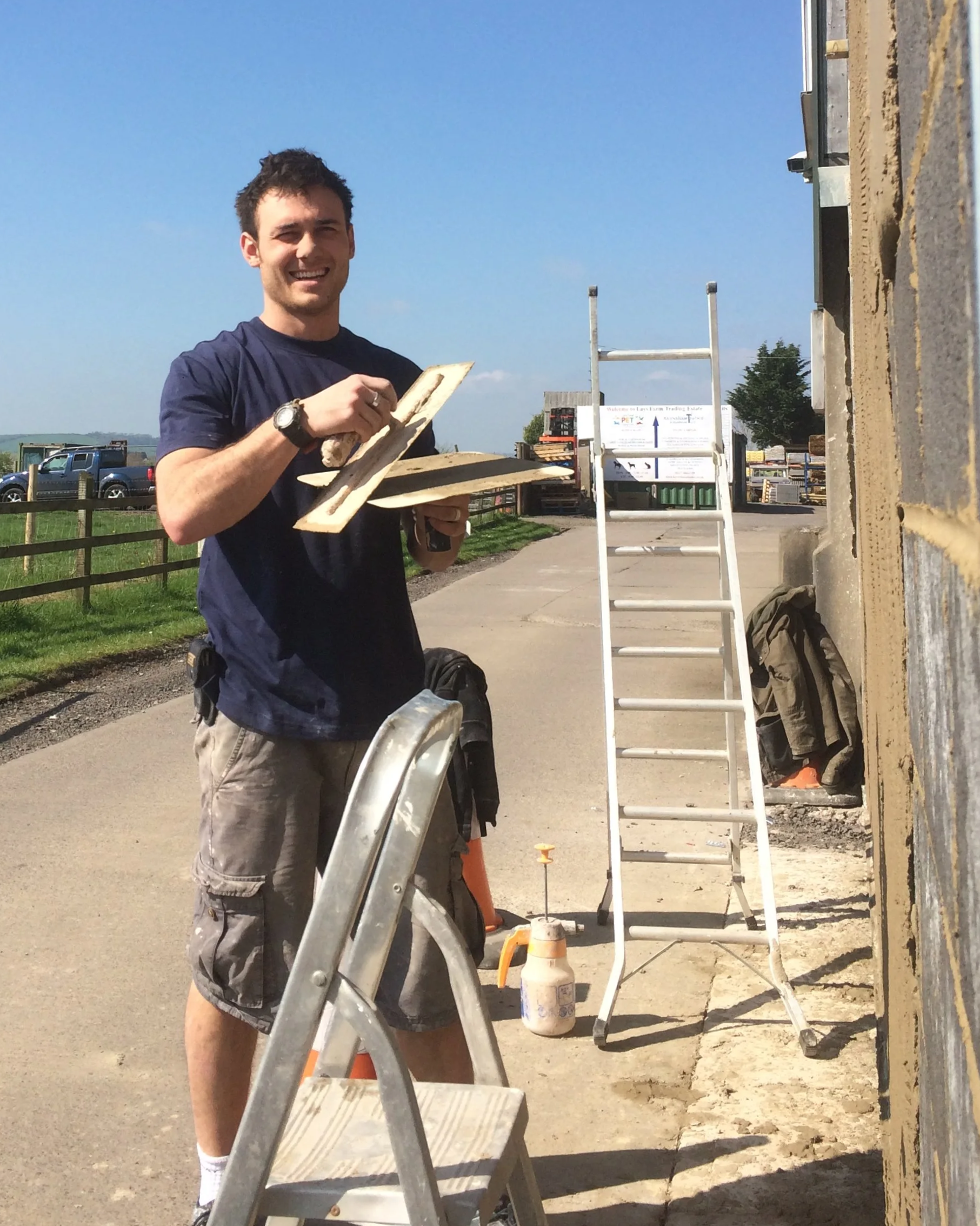 A man with a watch smiling and holding a wooden piece stands outdoors next to a ladder, during a sunny day, with construction materials and a building wall behind him.