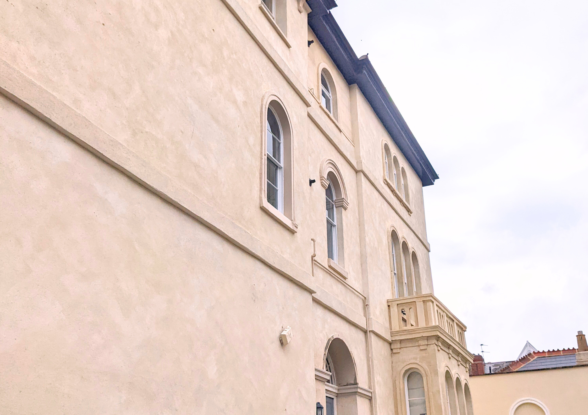 Front view of a cream-colored, multi-story residential building with arched windows, decorative moldings, and a balcony with balustrades, under a cloudy sky.