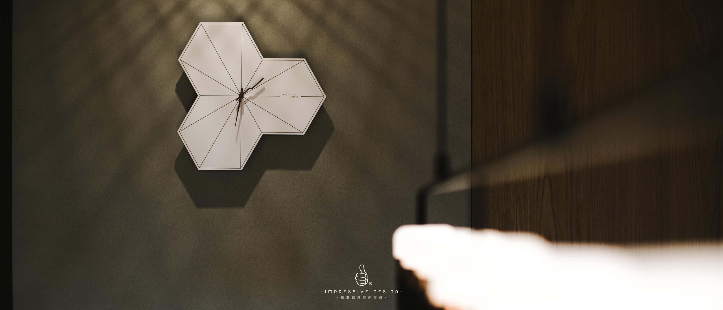 A hexagonal wall clock with black hands, mounted on a dark wall, casting a shadow. A part of a hanging lamp is visible in the foreground on the right. At the bottom, there is a logo and text that reads 'Impressive Design' with an icon of a hand givin