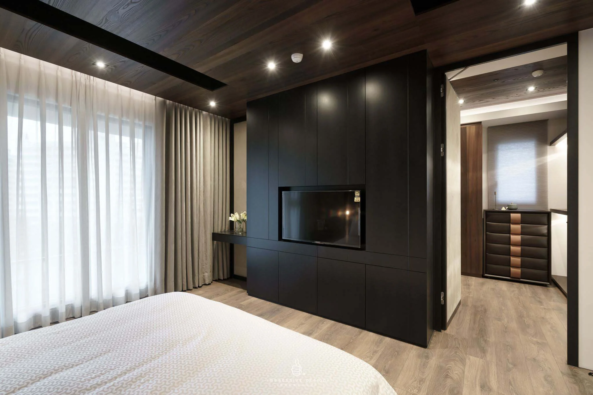 Modern bedroom with wooden ceiling, black wall unit with TV, white bed, large window with sheer and blackout curtains, and a glimpse of a dresser in the hallway.