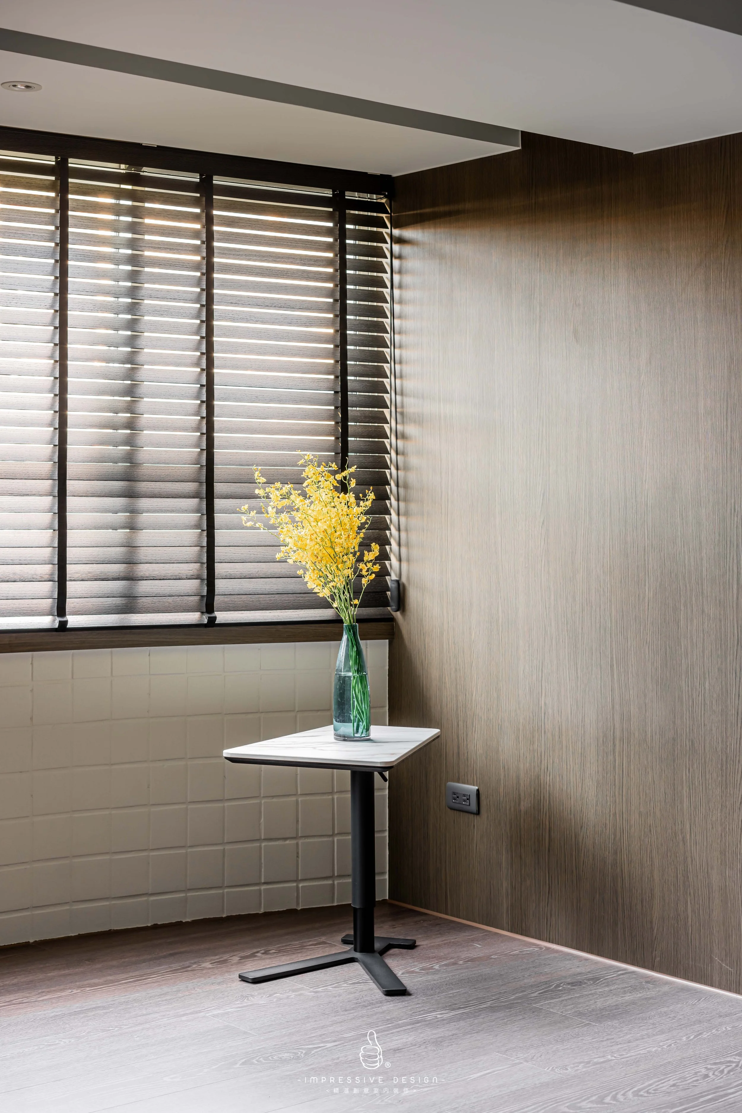 A room corner with a small side table holding a tall glass vase with yellow flowers, next to a window with horizontal blinds, and a textured beige wall.