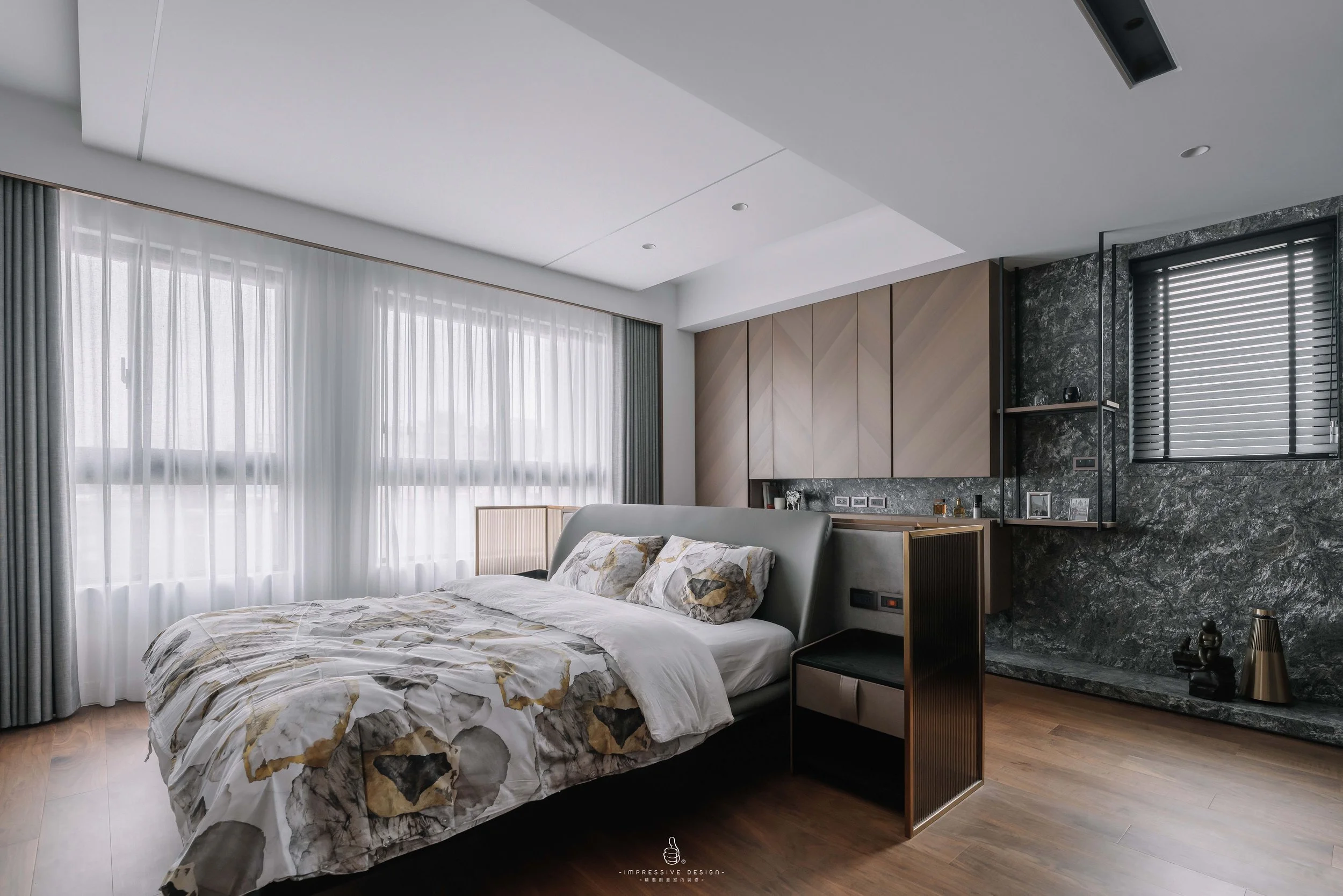 A modern bedroom with a large bed, patterned bedding, and large windows with sheer curtains, dark wood flooring, and contemporary decor.