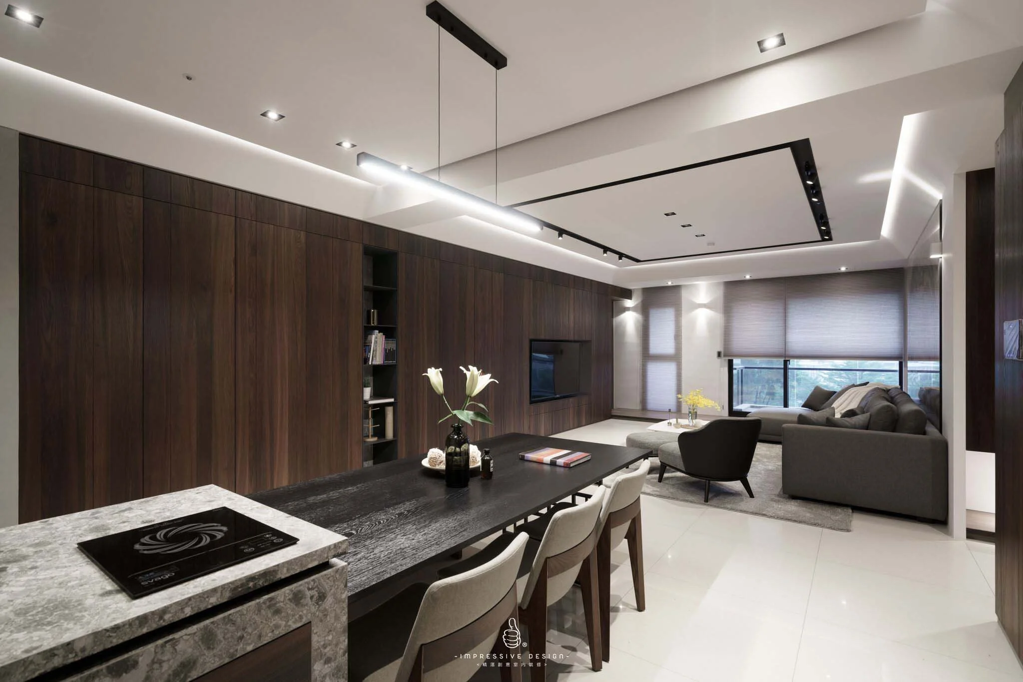 Modern living room and kitchen with dark wood wall, flat-screen TV, gray sectional sofa, black armchair, large windows with blinds, dining table with white chairs, flower vase, and minimalist ceiling lighting.