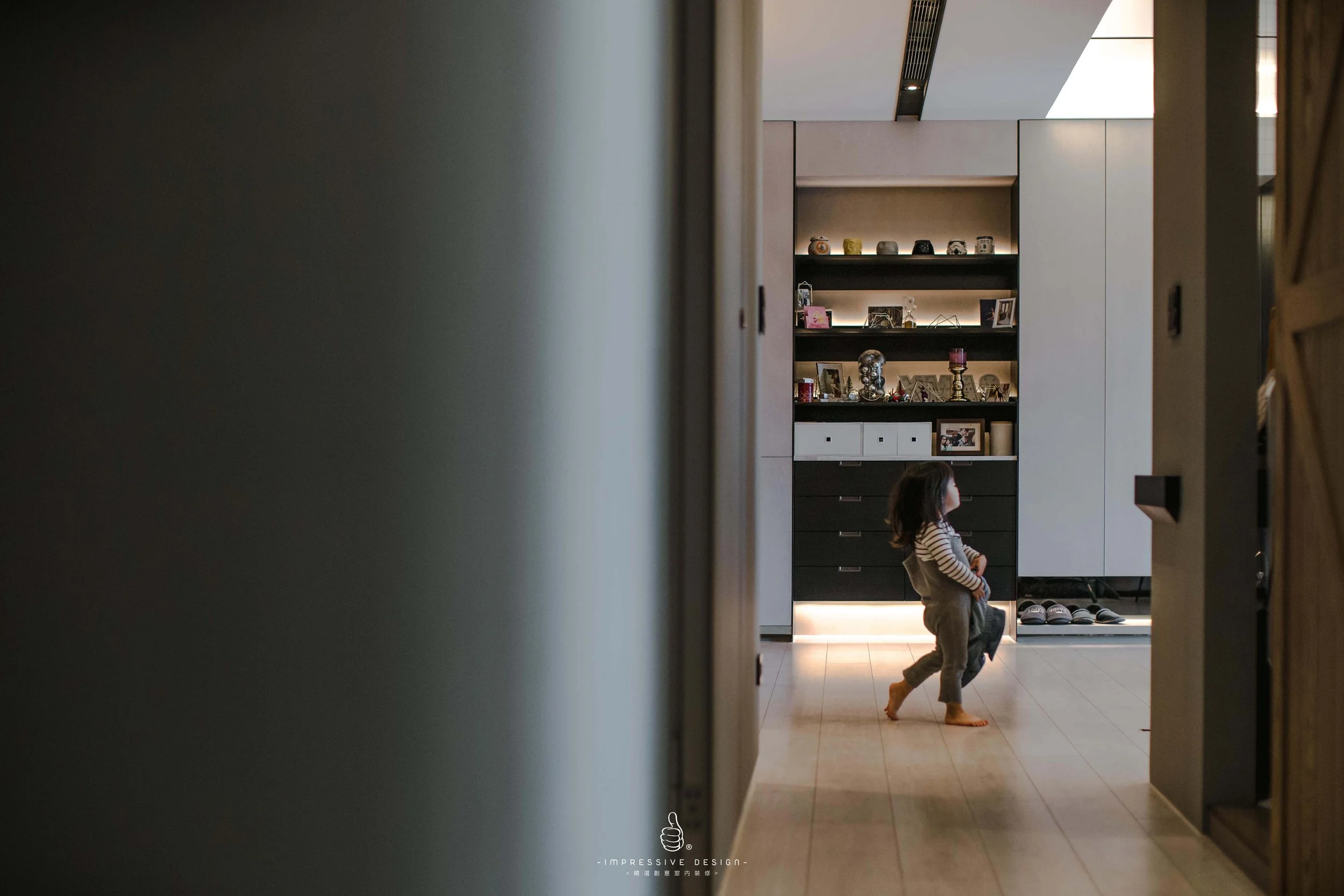 A young child playing barefoot in a modern apartment, seen through a doorway with shelves filled with decorative items and framed photos in the background.