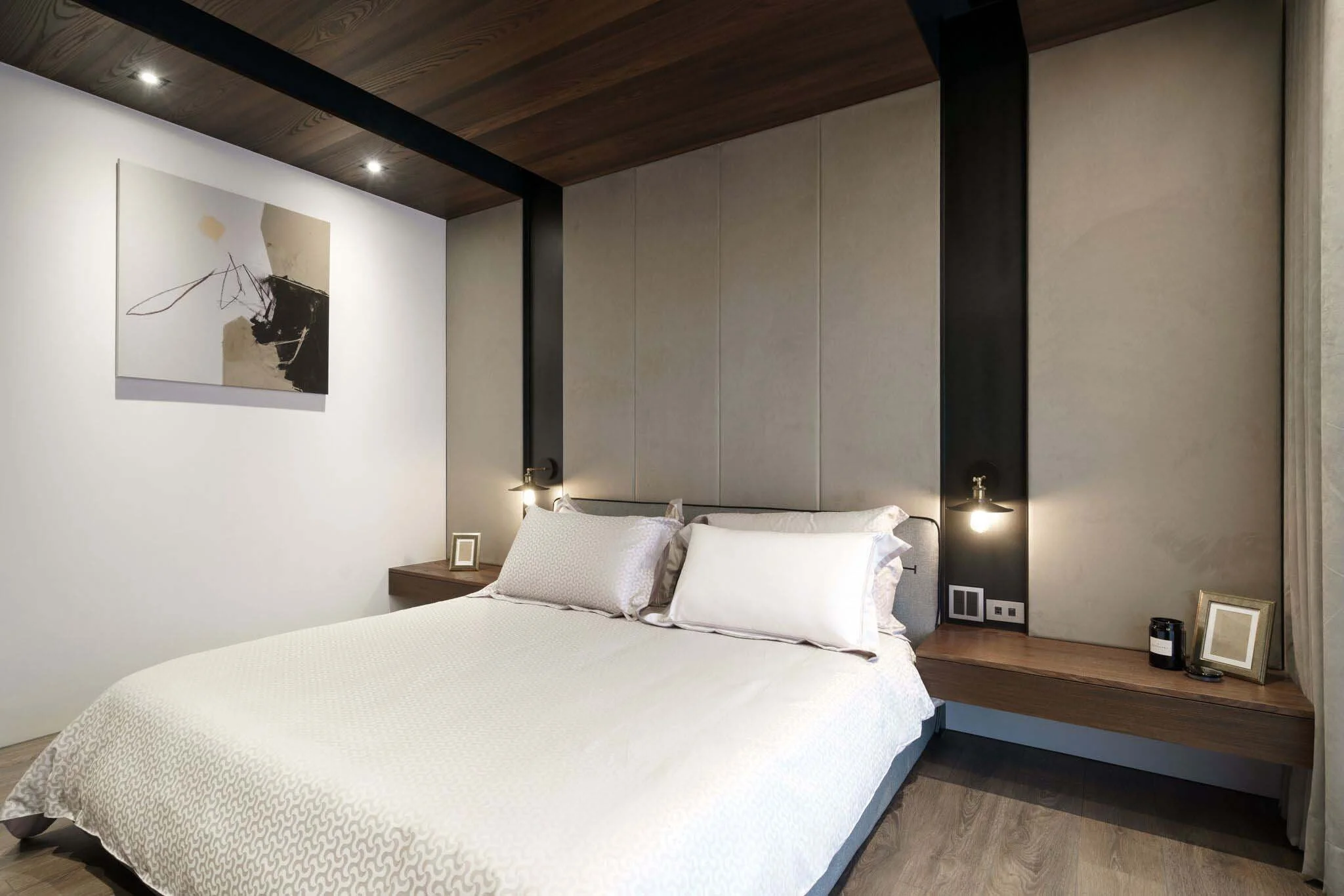 Modern bedroom with a large bed, white bedding, and pillows. Floating wooden nightstands on either side with small picture frames, a candle, and decorative items. Wall-mounted lamps and a large abstract artwork on the wall. Soft lighting creates a co