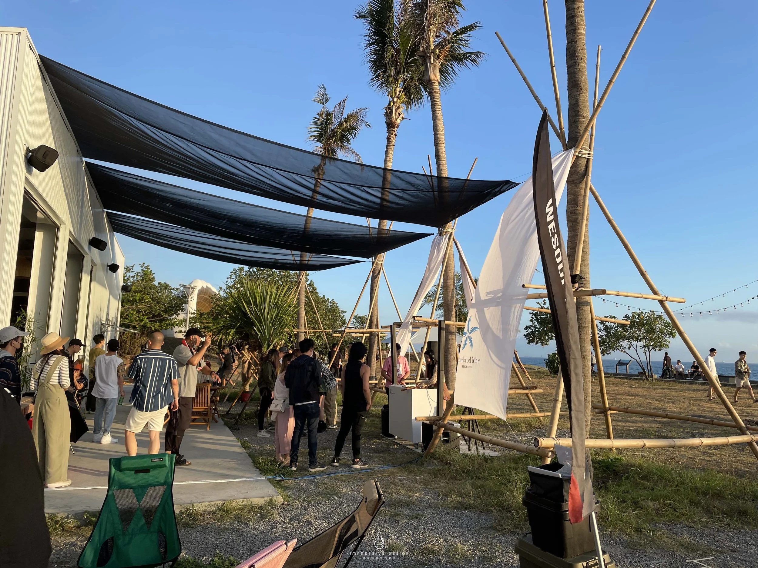 Group of people gathered outside near a building with black fabric sun shades, set on a grassy area with palm trees and a view of the ocean in the background.