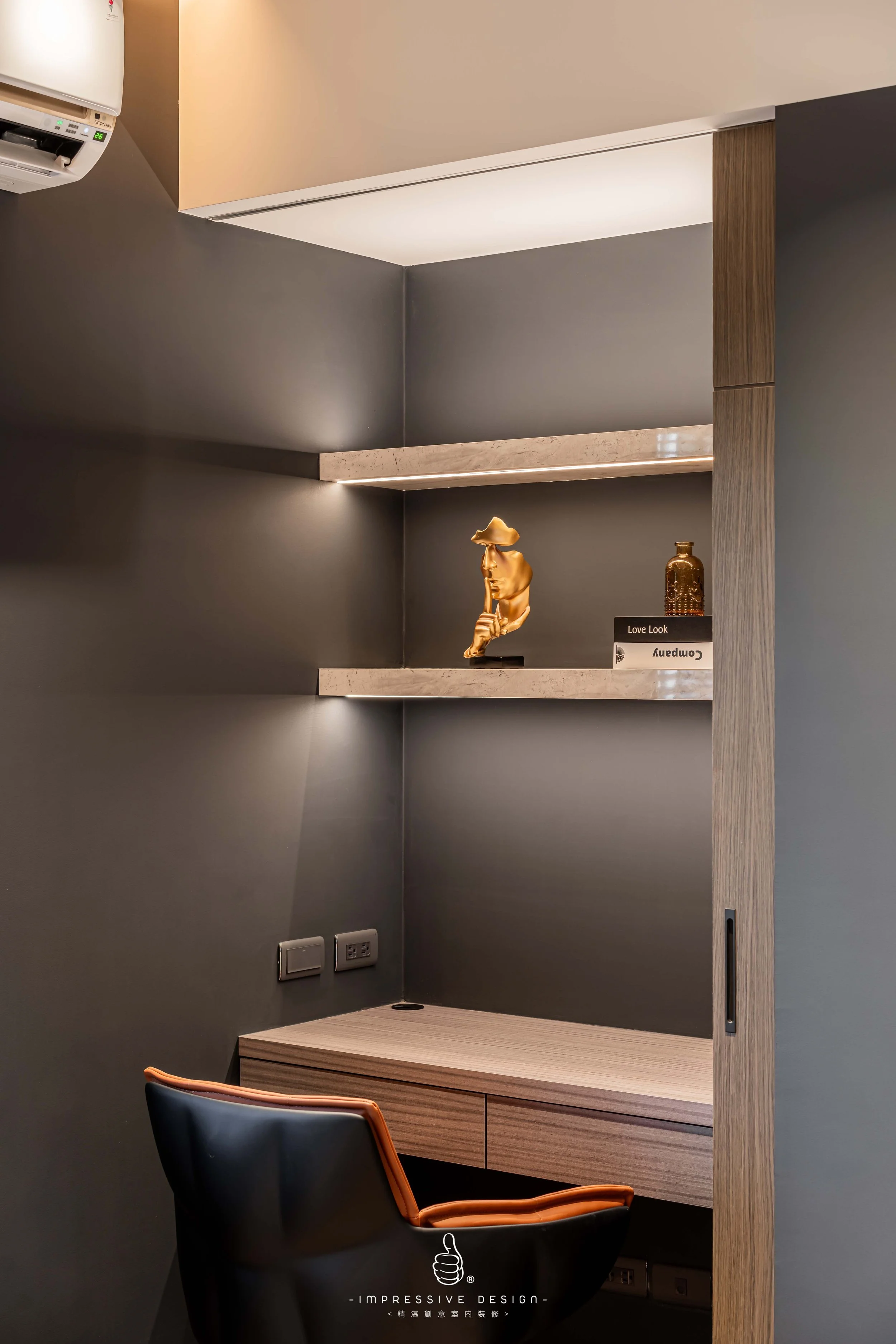 A modern workspace with a dark gray wall, wooden desk, and two floating shelves featuring decorative items. An office chair with black seat and orange trim is in front of the desk, with electrical outlets on the wall.