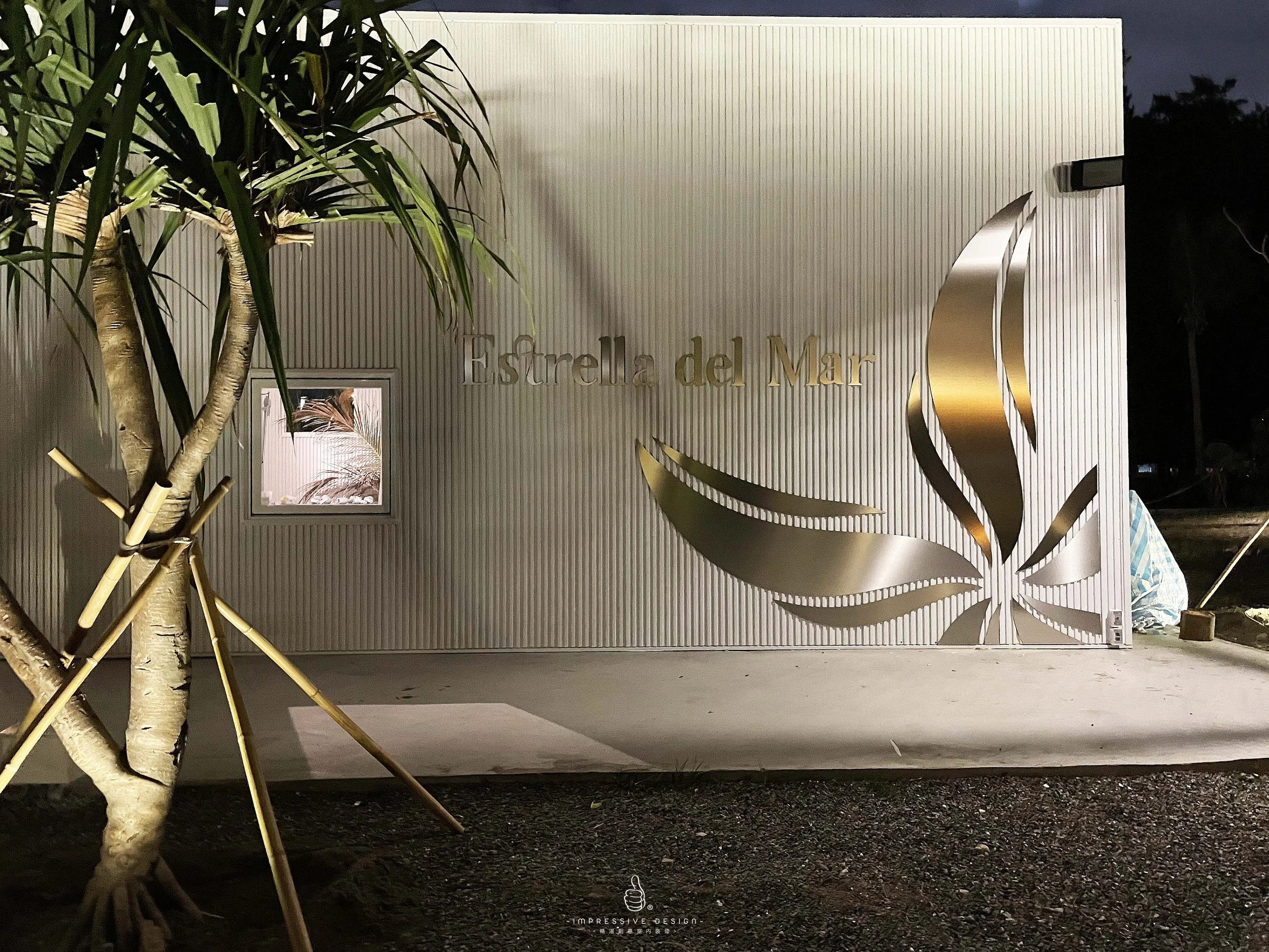 Nighttime view of a building with a white ribbed exterior wall, a gold logo of a stylized leaf, and the words 'Estrella del Mar' on the wall. There is a small window with palm leaves inside, and a large plant on the left side with a tree trunk and se