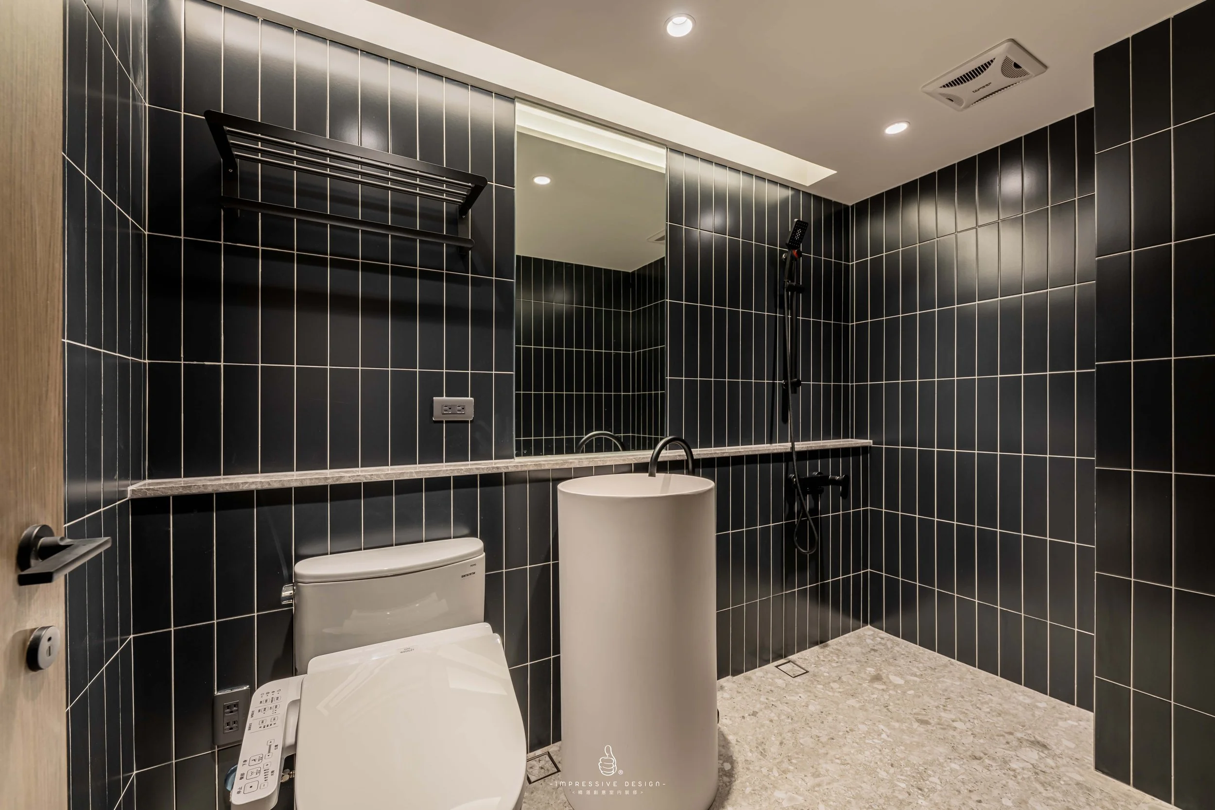 Modern bathroom with black tiled walls, a white toilet with electronic bidet, a white pedestal sink, a large mirror, a black towel rack, and a shower with a black showerhead and handheld shower wand.