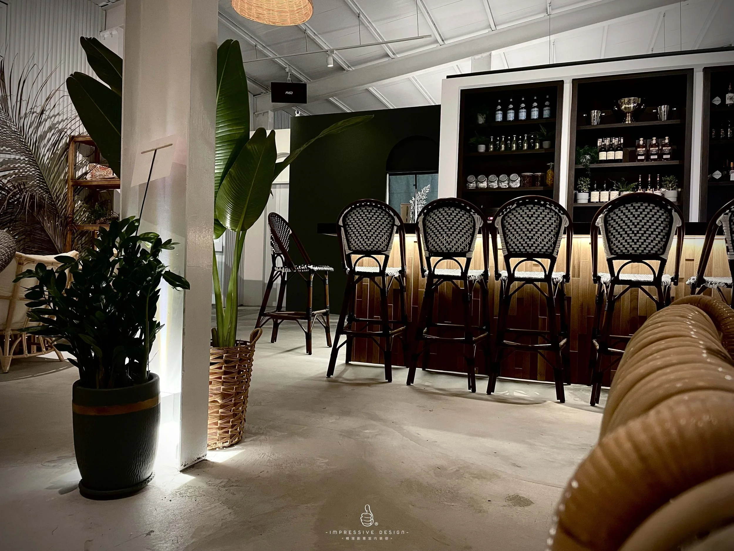 Interior of a cozy cafe or bar with bar stools lined up in front of a back bar shelf filled with bottles and jars, surrounded by potted plants, wicker furniture, and warm lighting.