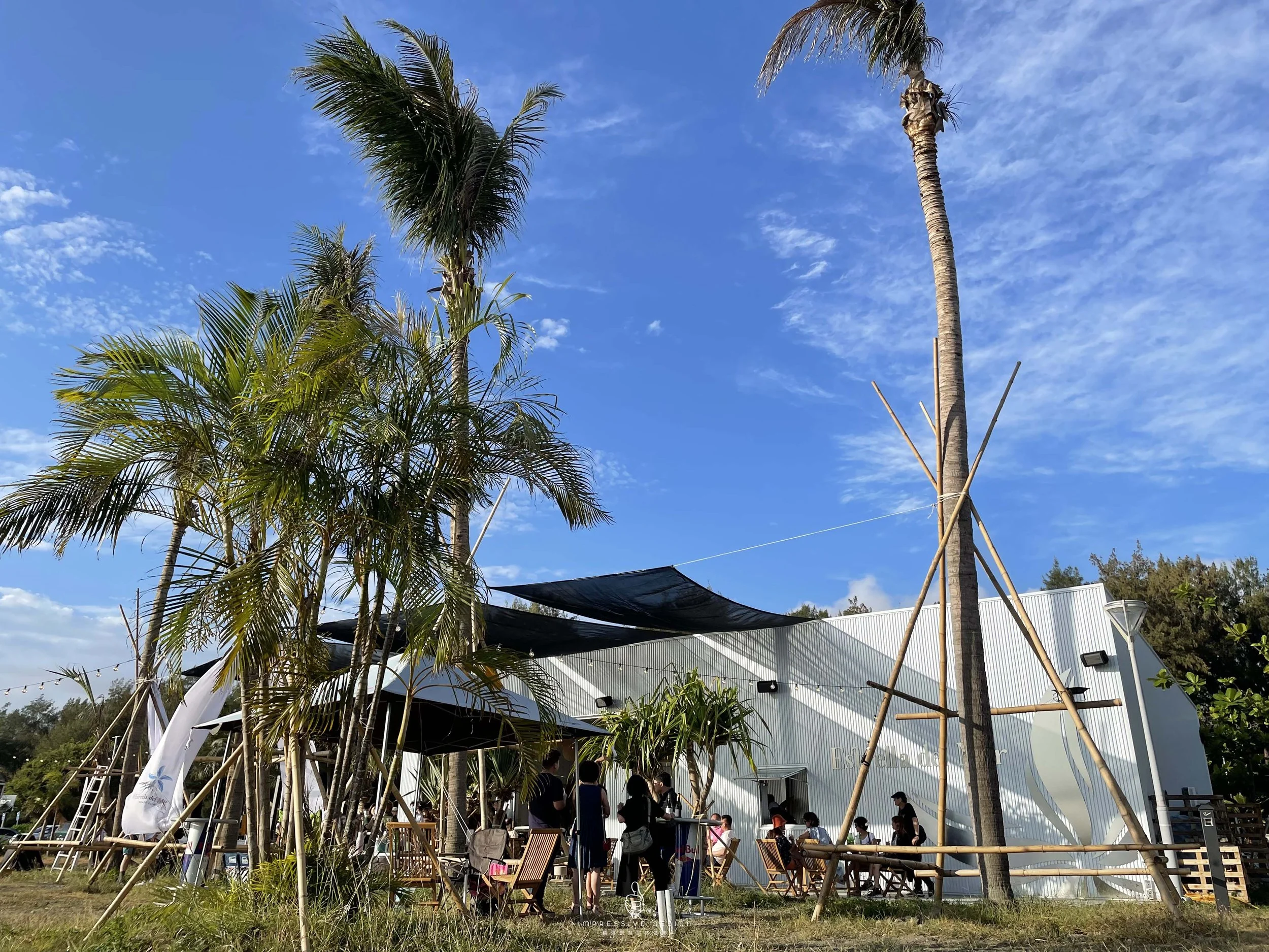 Outdoor scene with palm trees, a white building, and people gathered around tables under shade, with a blue sky and some clouds.