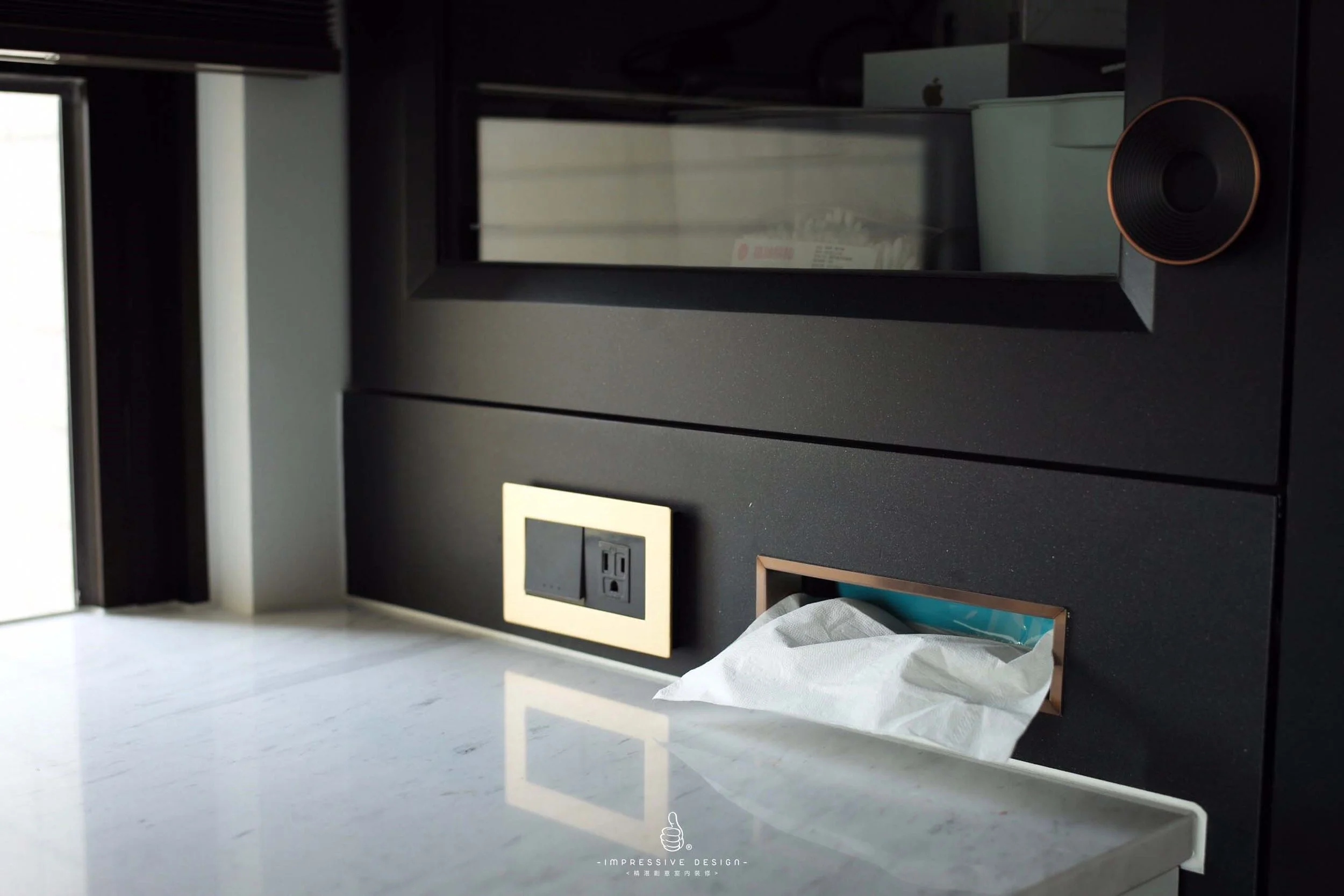 Close-up of a modern black wall with electrical outlets, a tissue dispenser with tissue, a black device on a black surface, and a mirrored cabinet above the outlets with a round black and copper speaker on the right.