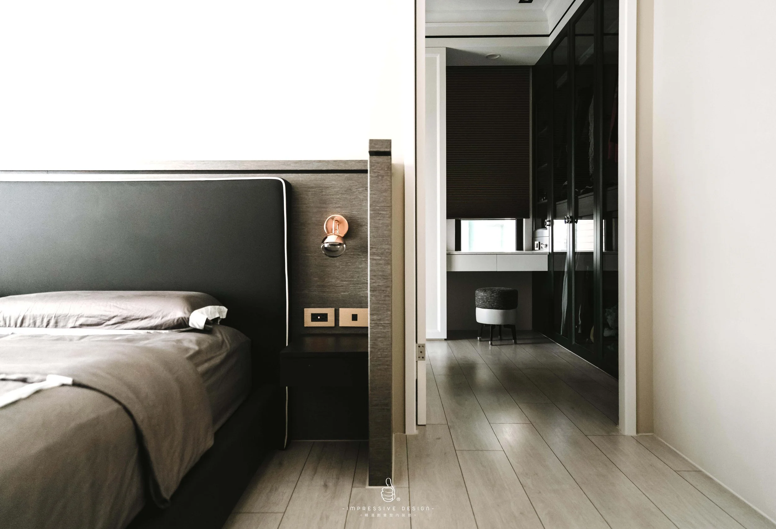 Modern bedroom with a large dark headboard, small nightstand, and light-colored bedding. Partial view of an adjoining room with a window, black cabinetry, and a small stool.