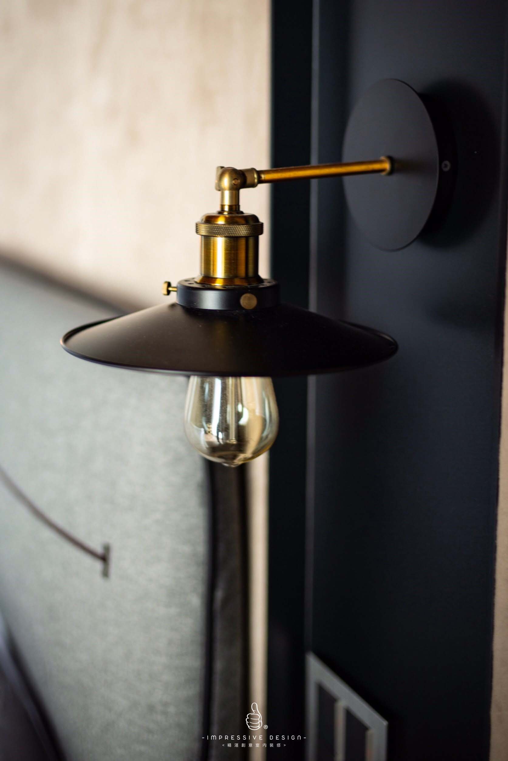 Close-up of a modern wall-mounted black and brass lamp with a glass bulb, set against a dark wall with a grey textured background.