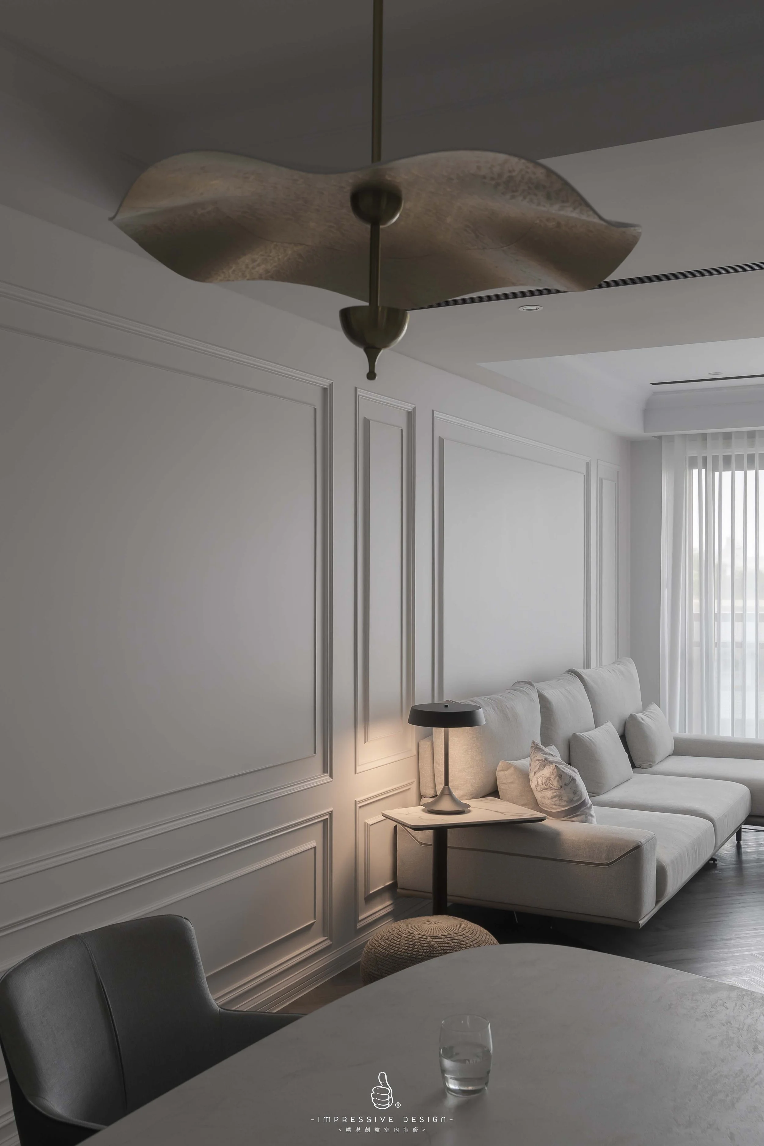 Living room with a white sofa against a wall with decorative paneling, a black table lamp on a side table, a small round woven ottoman, a window with vertical blinds, and a dining table with a glass of water in the foreground.