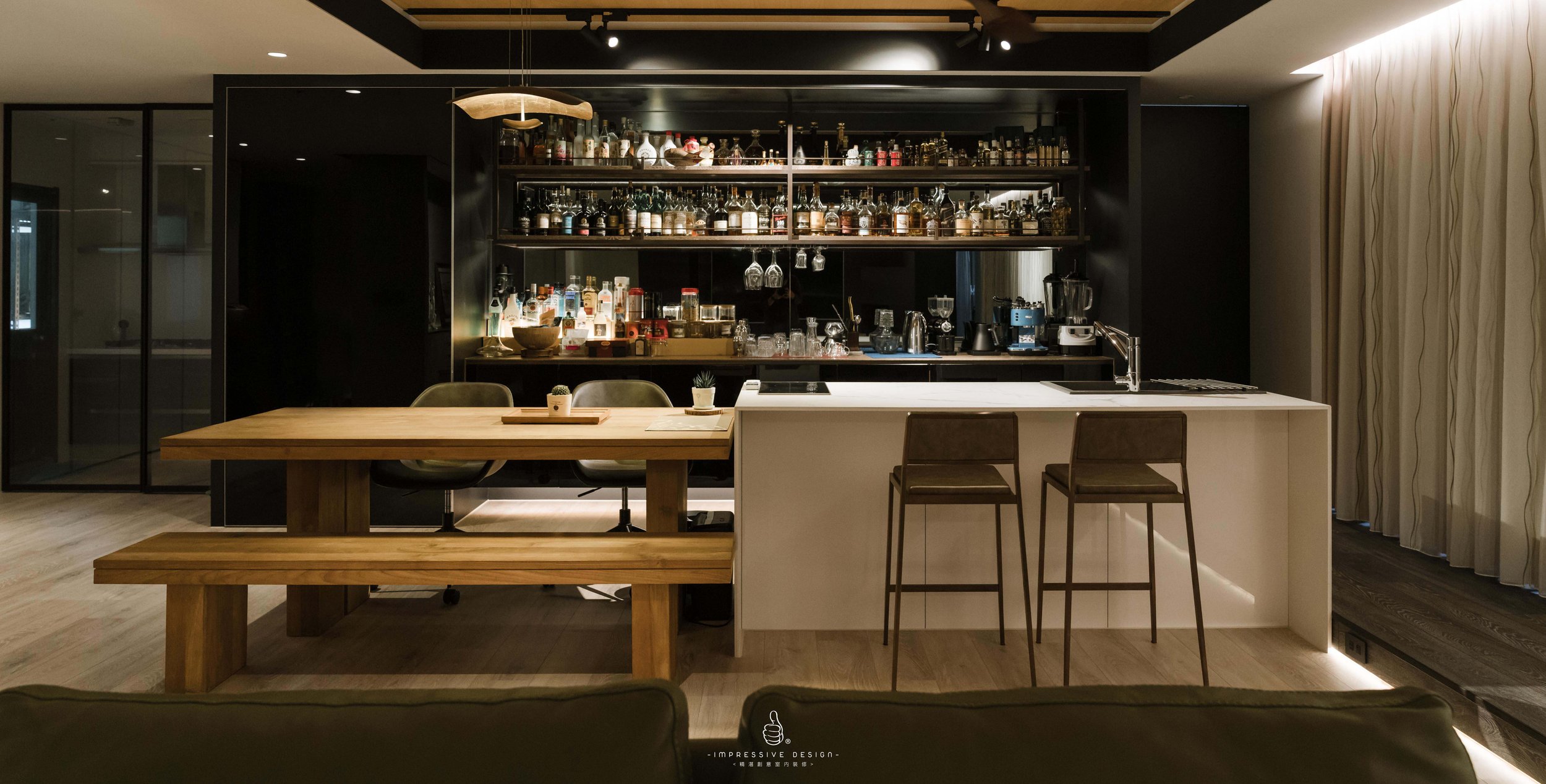 Modern home bar with black wall-mounted shelves filled with bottles, glasses, and barware. A white kitchen island with seating and a wooden dining table with benches in the foreground. Low lighting creates a cozy ambiance.