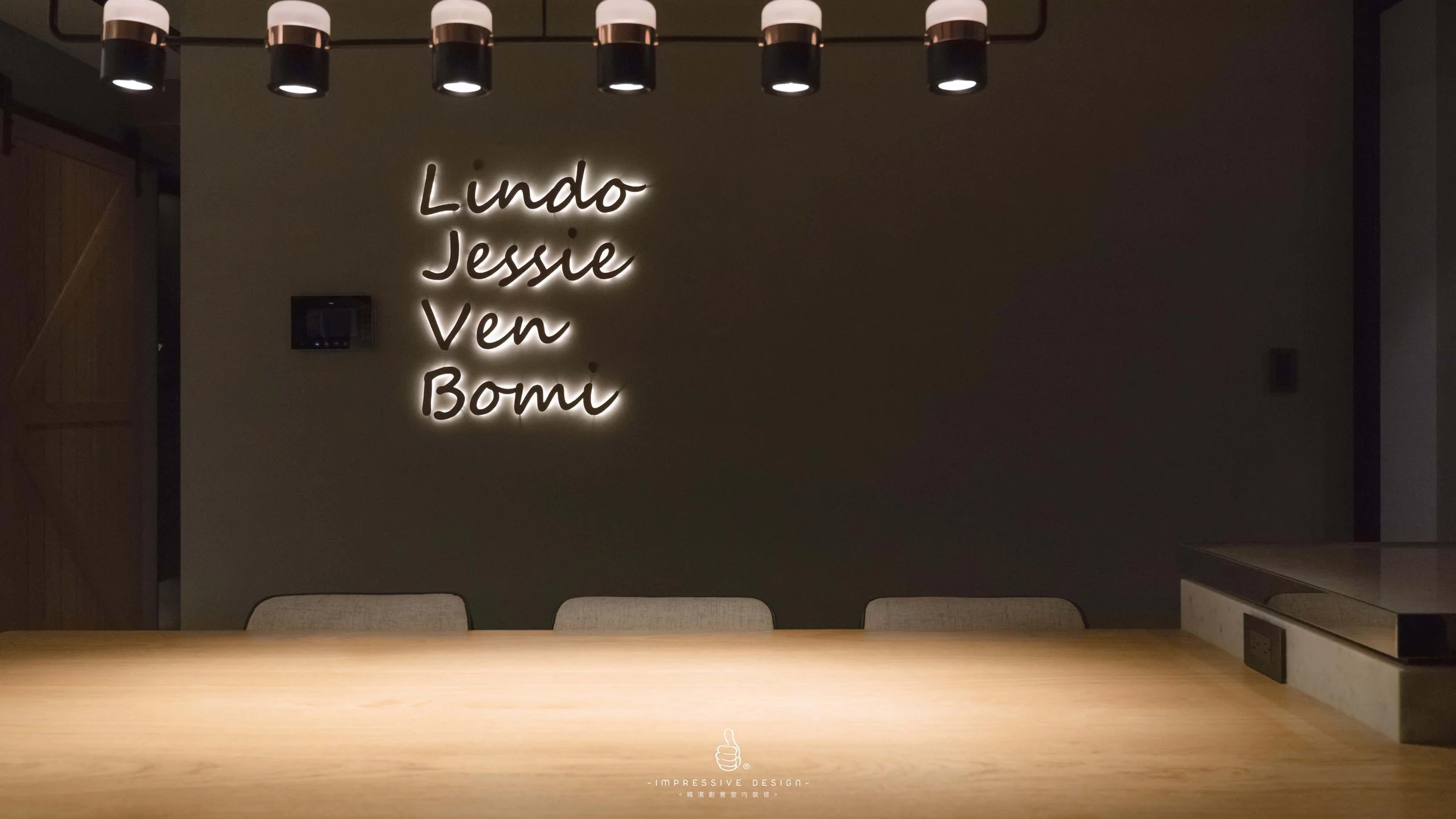 Interior of a modern room with a neon sign on the wall displaying the names Lindo, Jessie, Ven, and Bomi, a wooden table, beige chairs, and black ceiling-mounted light fixtures.
