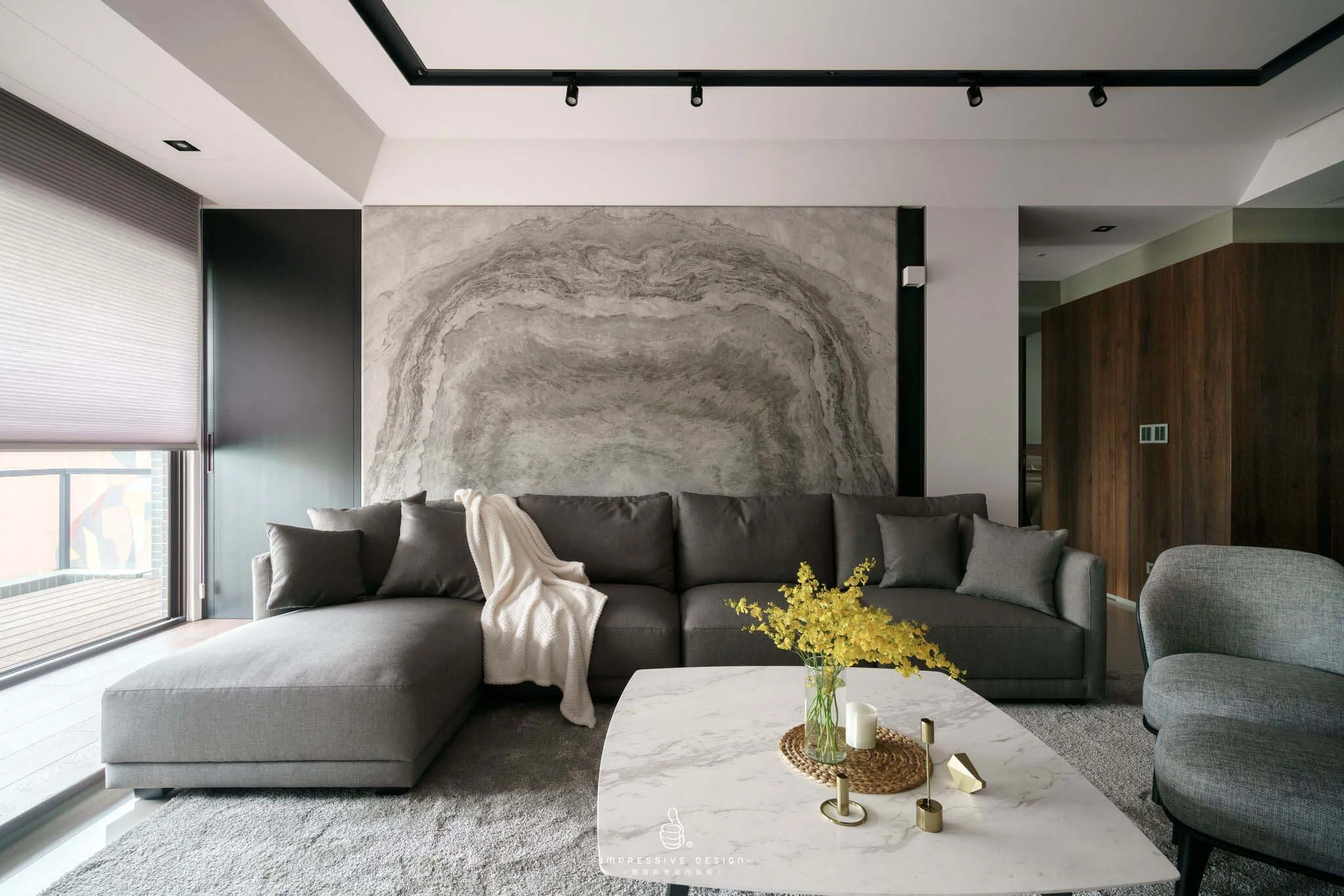 Modern living room with a gray sectional sofa, a marble coffee table with decorative items, a large abstract wall art behind the sofa, and a sliding glass door with blinds to the left, letting in natural light.