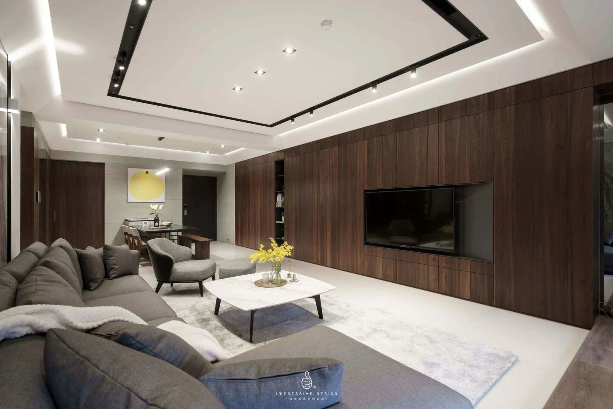 Modern living room with a large gray sectional sofa, two gray armchairs, a white marble coffee table with yellow flowers, a wall-mounted flat-screen TV, wooden wall paneling, and a dining area in the background with a yellow hanging light.