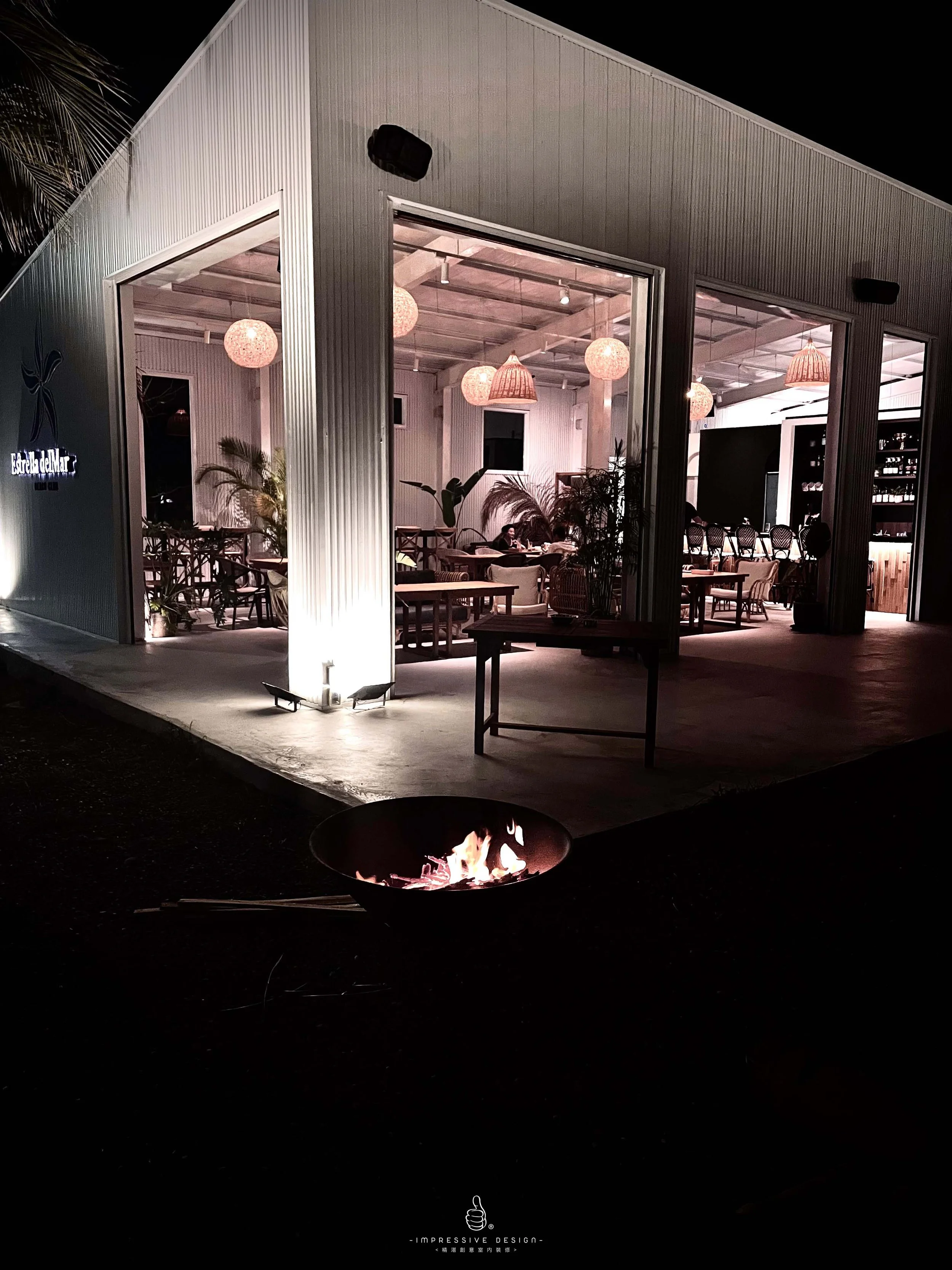 A modern, illuminated restaurant with large glass windows and tropical plants inside. Outside, there is a fire pit with flames, set on a dark outdoor area at night.