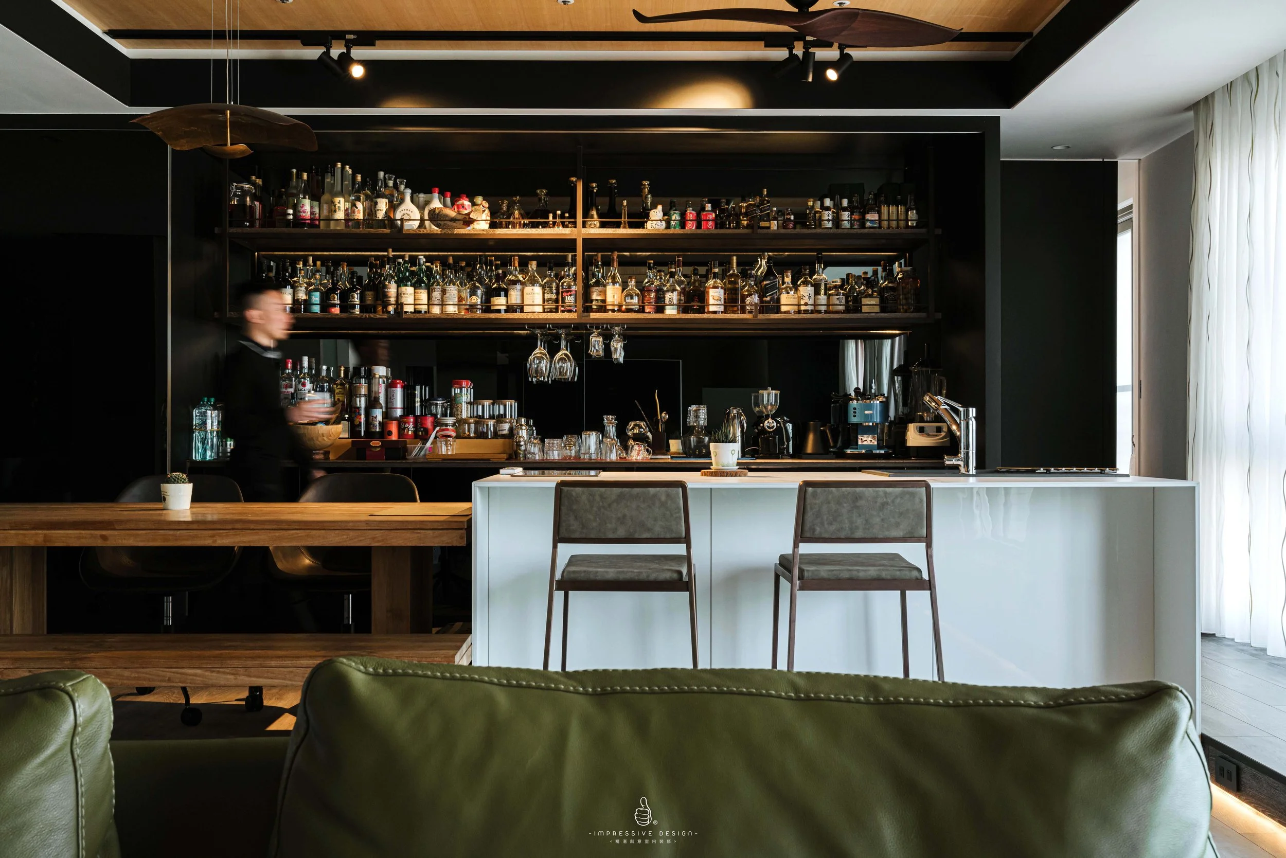 Modern bar area with a black wall, wooden shelves stocked with various liquor bottles, a white counter with two barstools, and a partially visible person's blurred motion on the left. There are green leather chairs in the foreground and a large windo