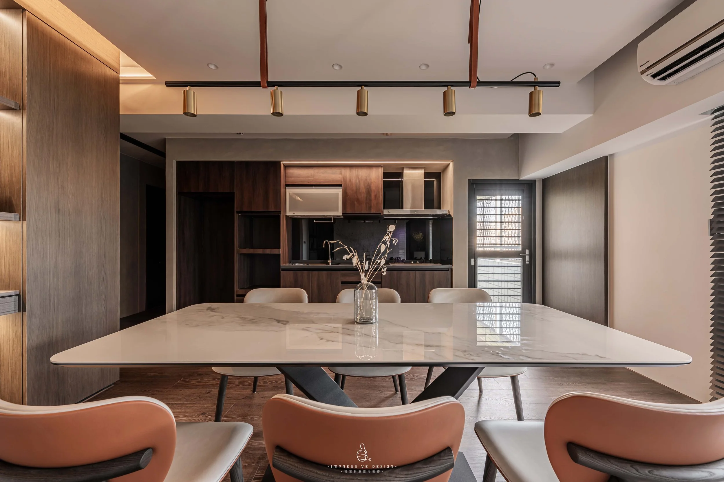 Modern dining area with a white marble table, beige chairs, a glass vase with dried flowers, wooden cabinets, a small kitchen with a stainless steel range hood, and a door leading outside.