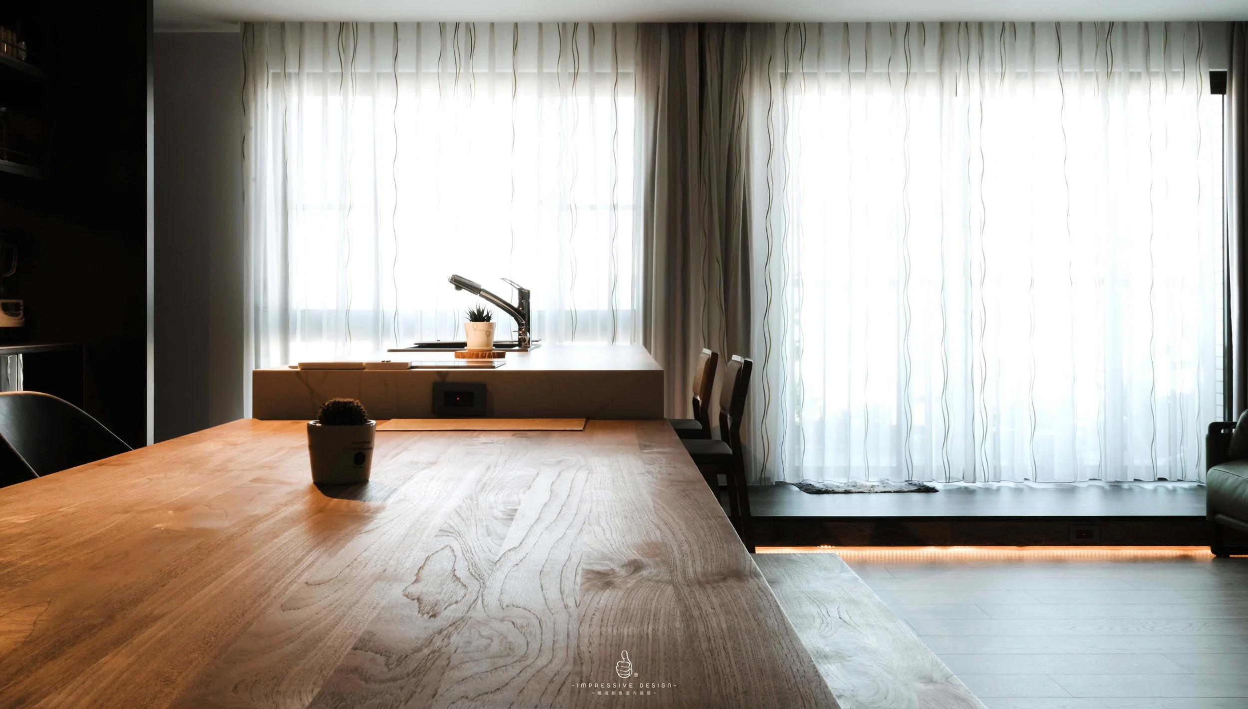 Interior view of a modern living space with a large wooden dining table, potted plants, and sheer white curtains letting in natural light.