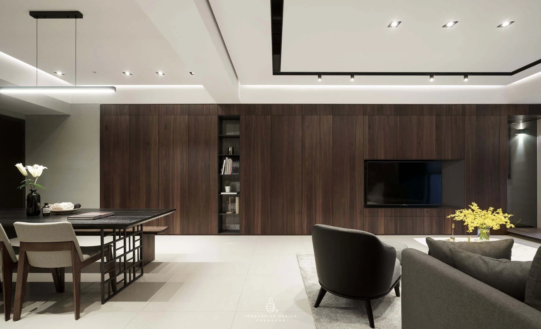 Modern living room with dark wood wall panels, a wall-mounted flat-screen TV, a black and beige dining table with chairs, a gray armchair, a sofa, and vases with flowers.