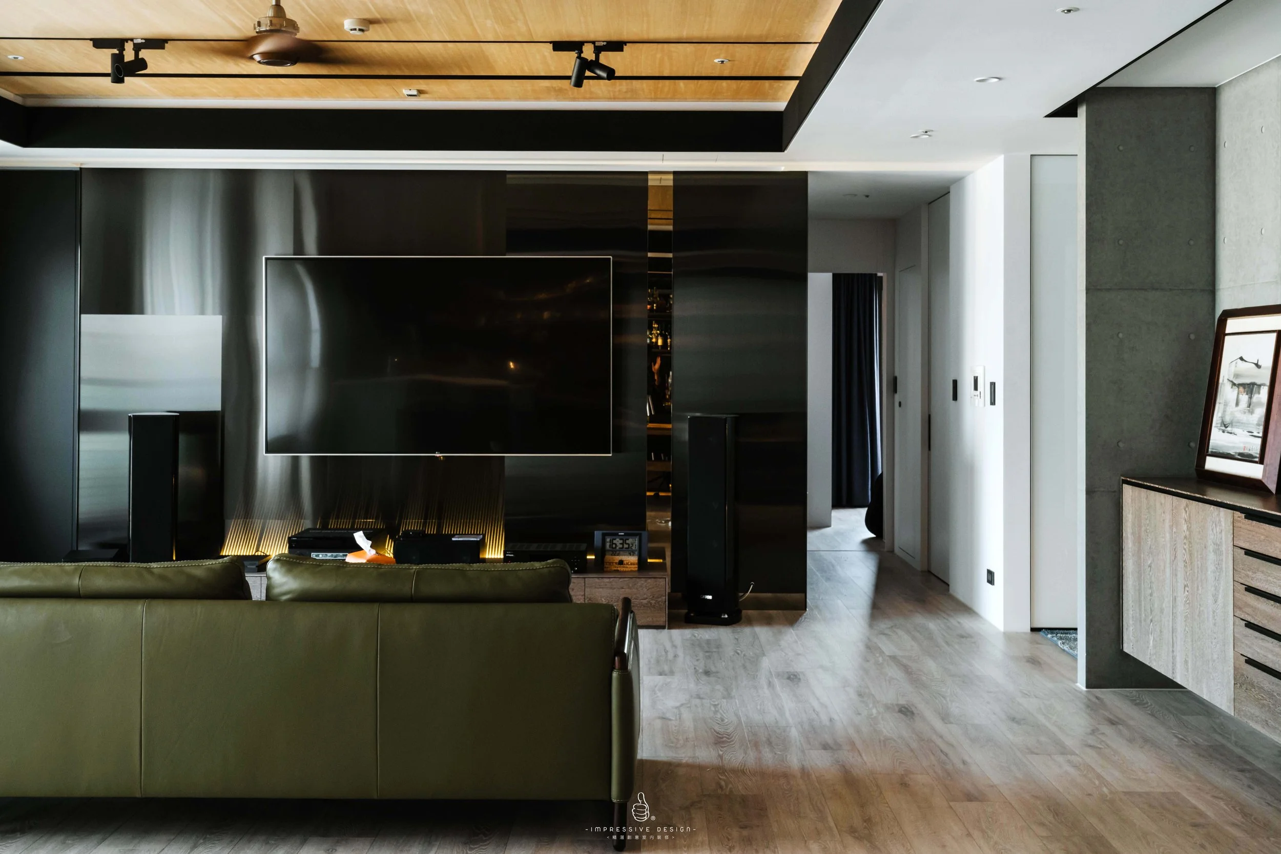 Modern living room with a large flat-screen TV on a black wall, olive green sofa, wooden flooring, and artwork on a wooden cabinet. Entryway with white and dark walls, and curtains in the background.