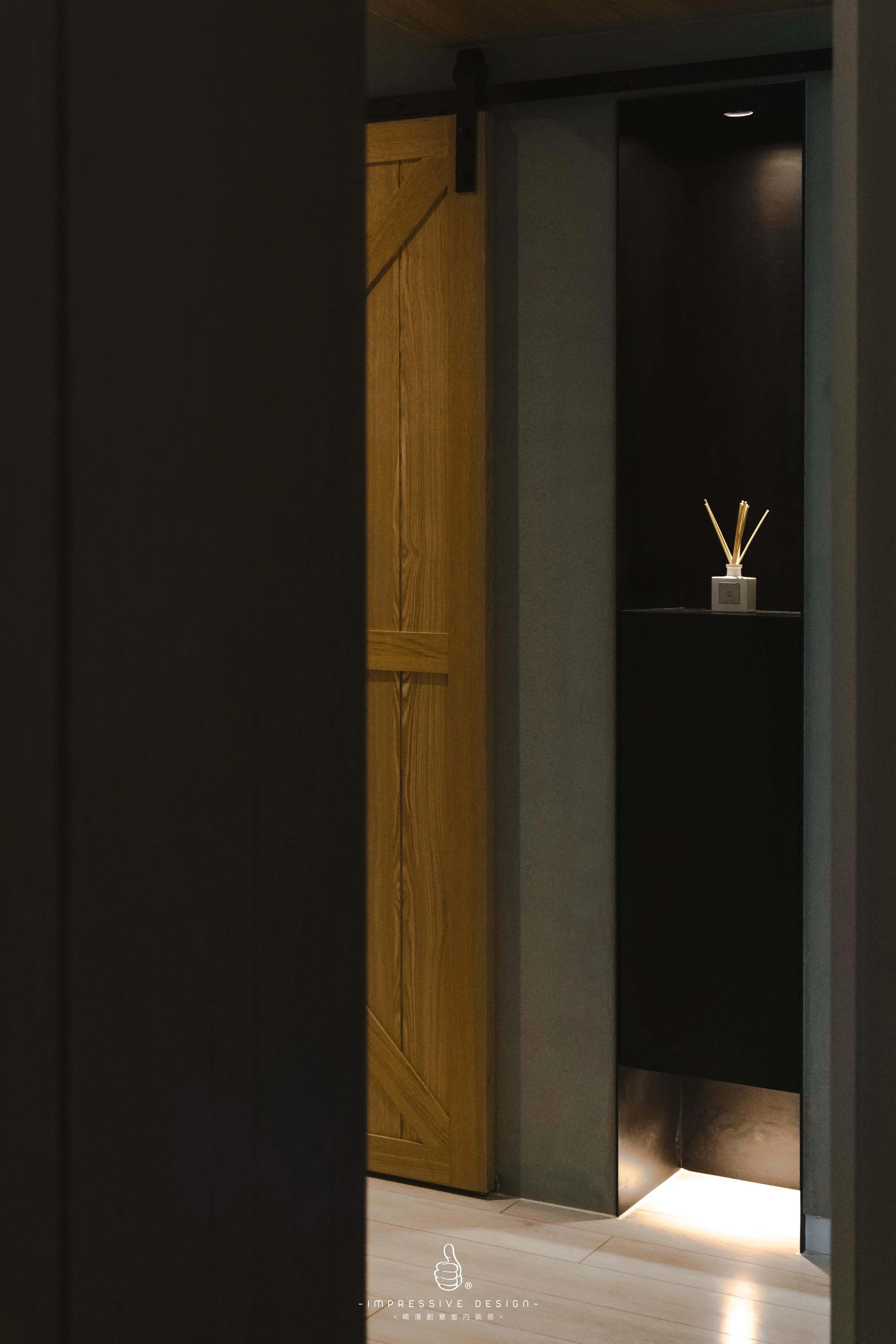 A partially open wooden door reveals a dimly lit hallway with a small black shelf holding a decorative object with reed sticks. The lower part of the scene shows light-colored wooden flooring and a logo on the floor that says 'Impressive Design'.