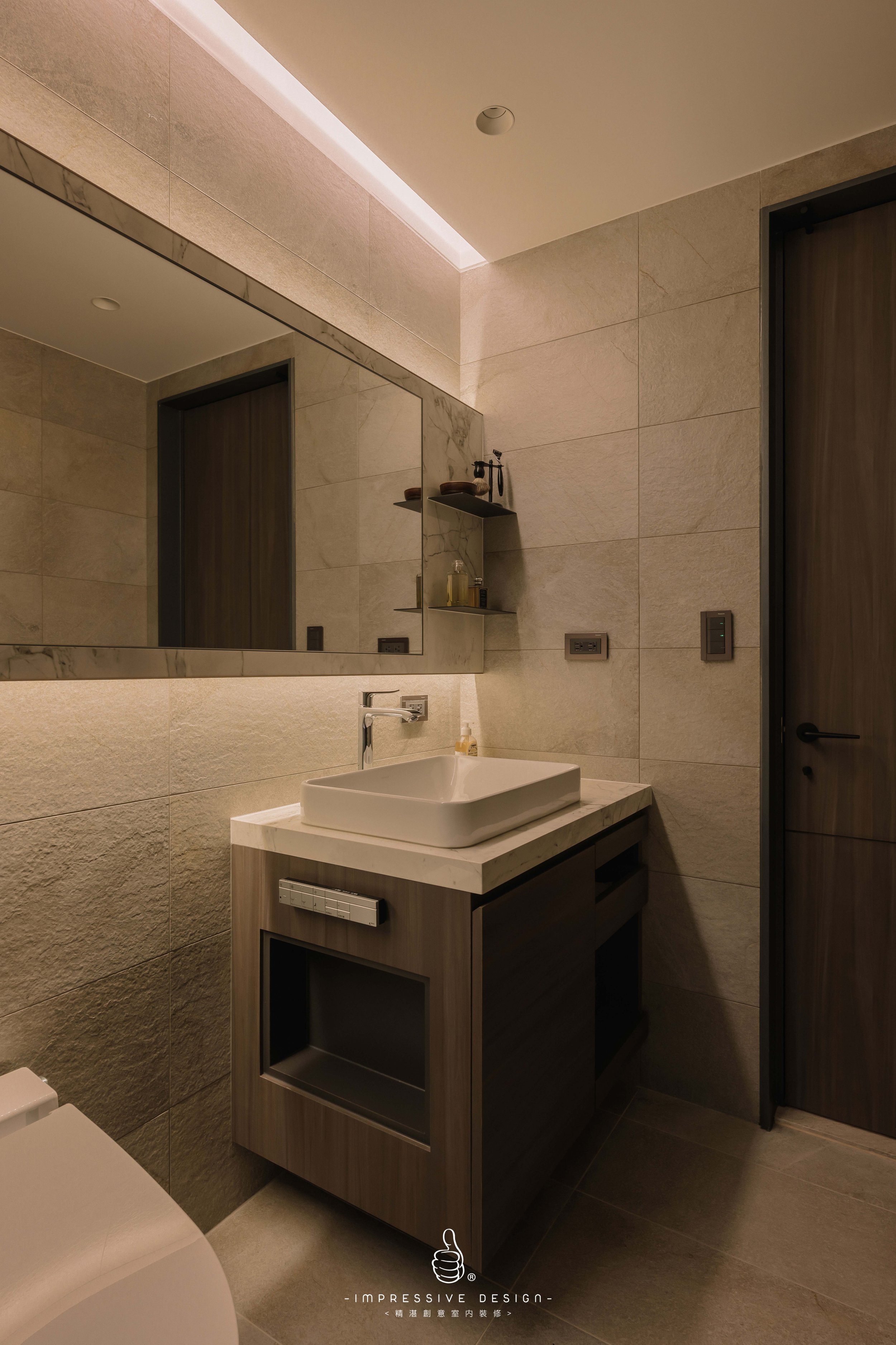 Modern bathroom with beige tiled walls and floor, a large rectangular mirror, a white vessel sink on a wooden vanity, a soap dispenser, a floating shelf with toiletries, and a wooden door.