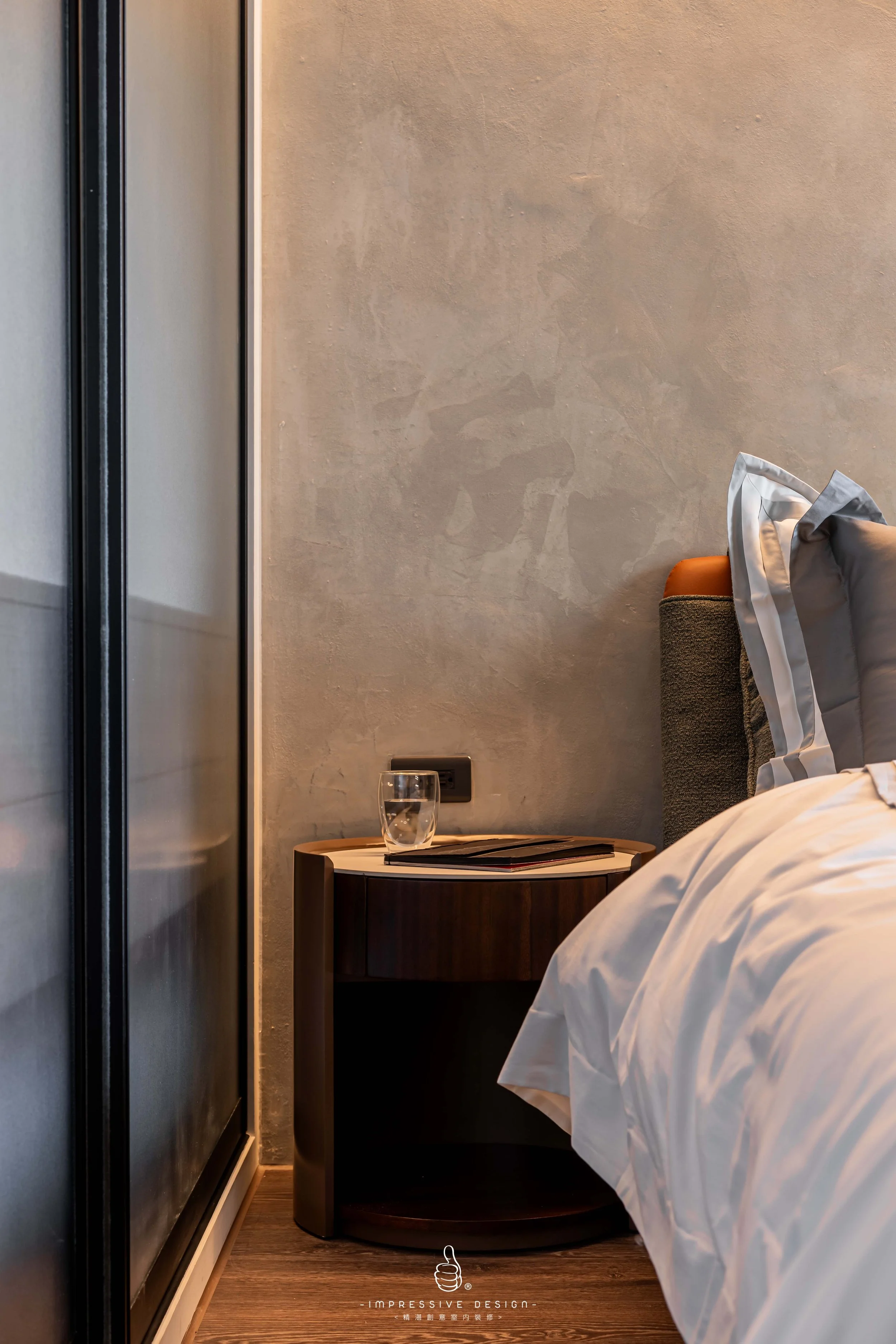 A bedroom with a bedside table holding a glass of water and magazines, a bed with pillows and white bedding, and a textured wall in the background.