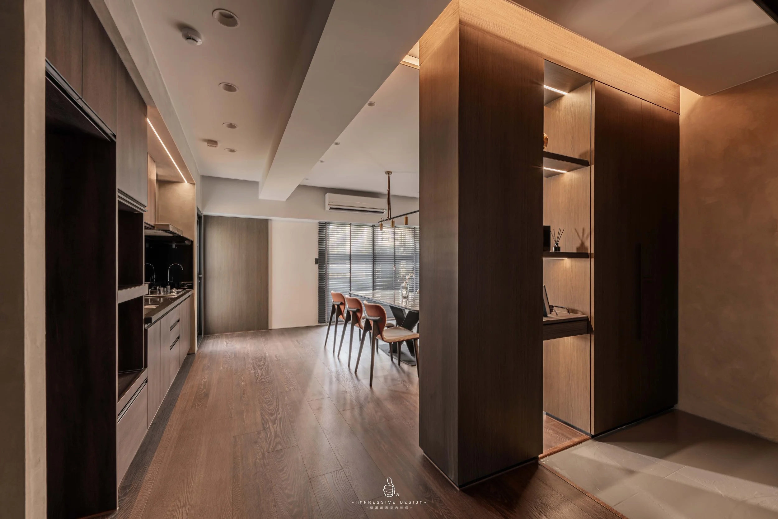 Modern dining area with a wooden dining table and four chairs near a window with blinds, adjacent to a kitchen with dark cabinetry, and a wooden partition with built-in shelves.