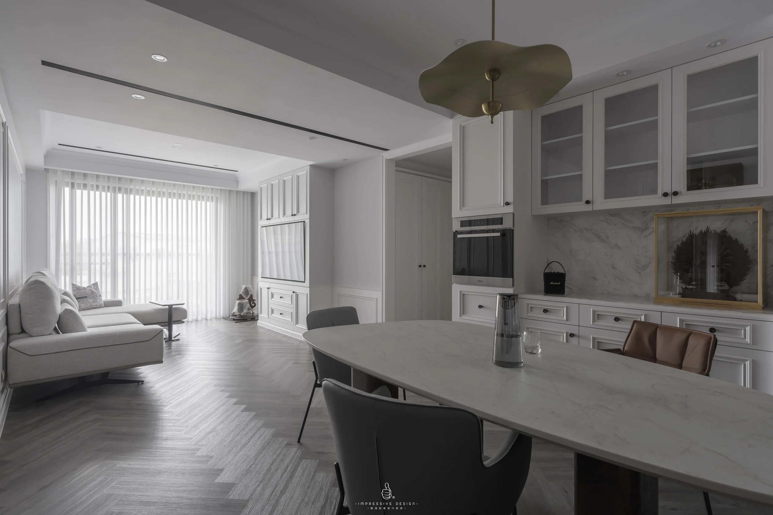 Modern living room and kitchen with white cabinetry, built-in oven, a marble countertop, a leather chair, a remote control on table, large window with vertical blinds, gray wooden floor, and contemporary light fixture.