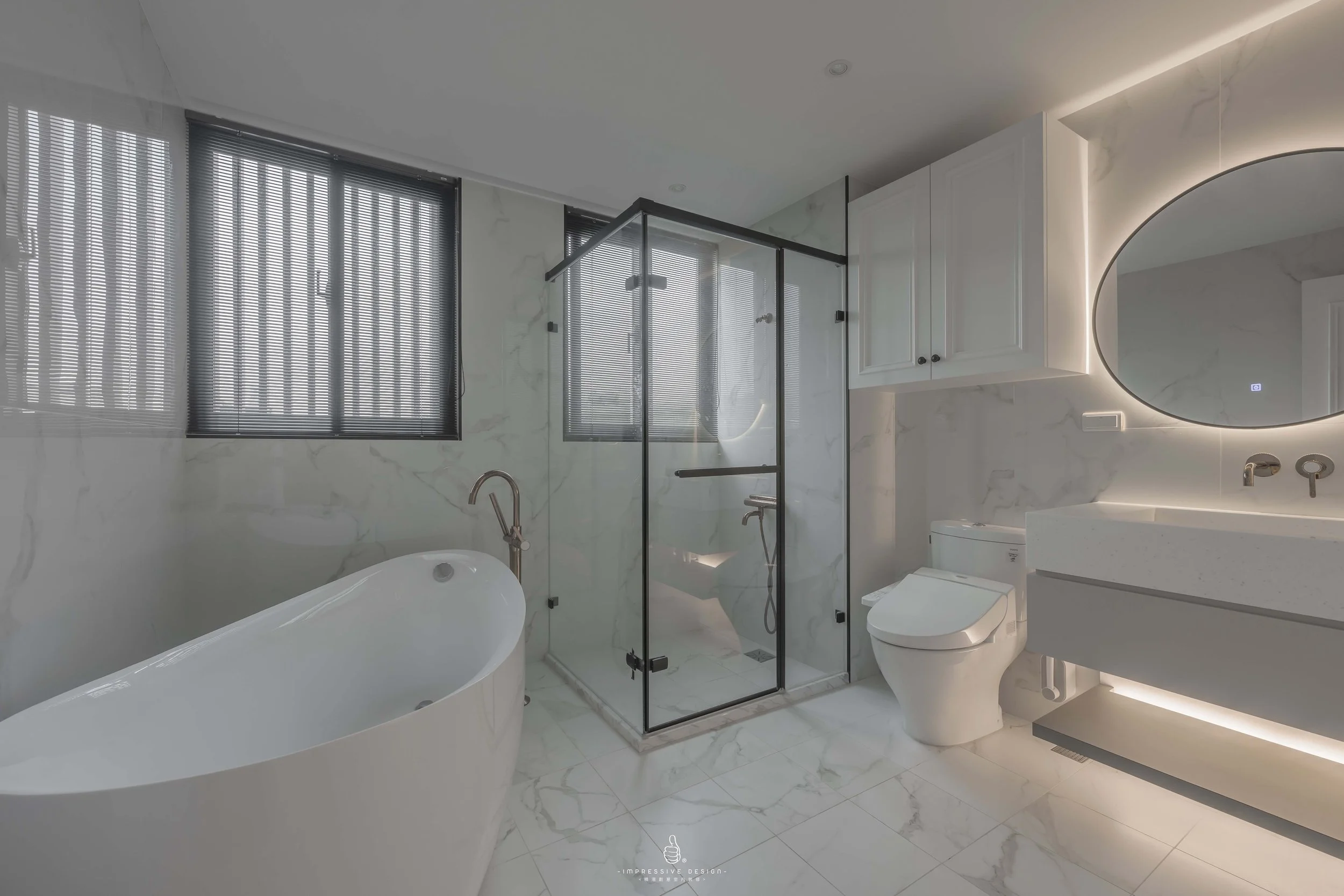 Modern bathroom with white marble walls and floor, a freestanding bathtub, a glass-enclosed shower, a wall-mounted toilet, a white vanity with a circular backlit mirror, and dark window blinds.