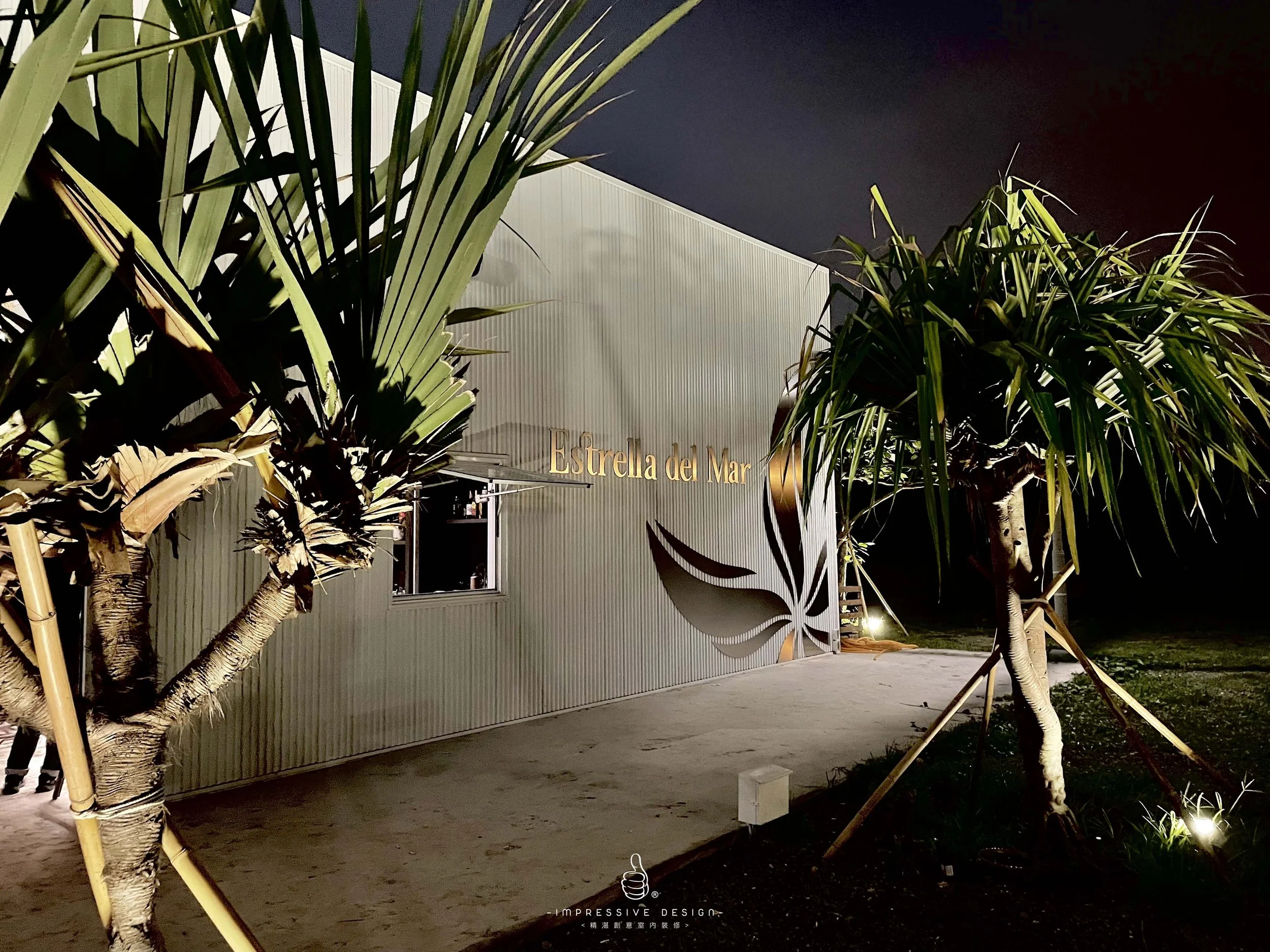 Nighttime view of a modern exterior building with gold lettering reading 'Estrela del Mar' and decorative metal elements resembling a leaf, with two palm trees in the foreground illuminated by lights.