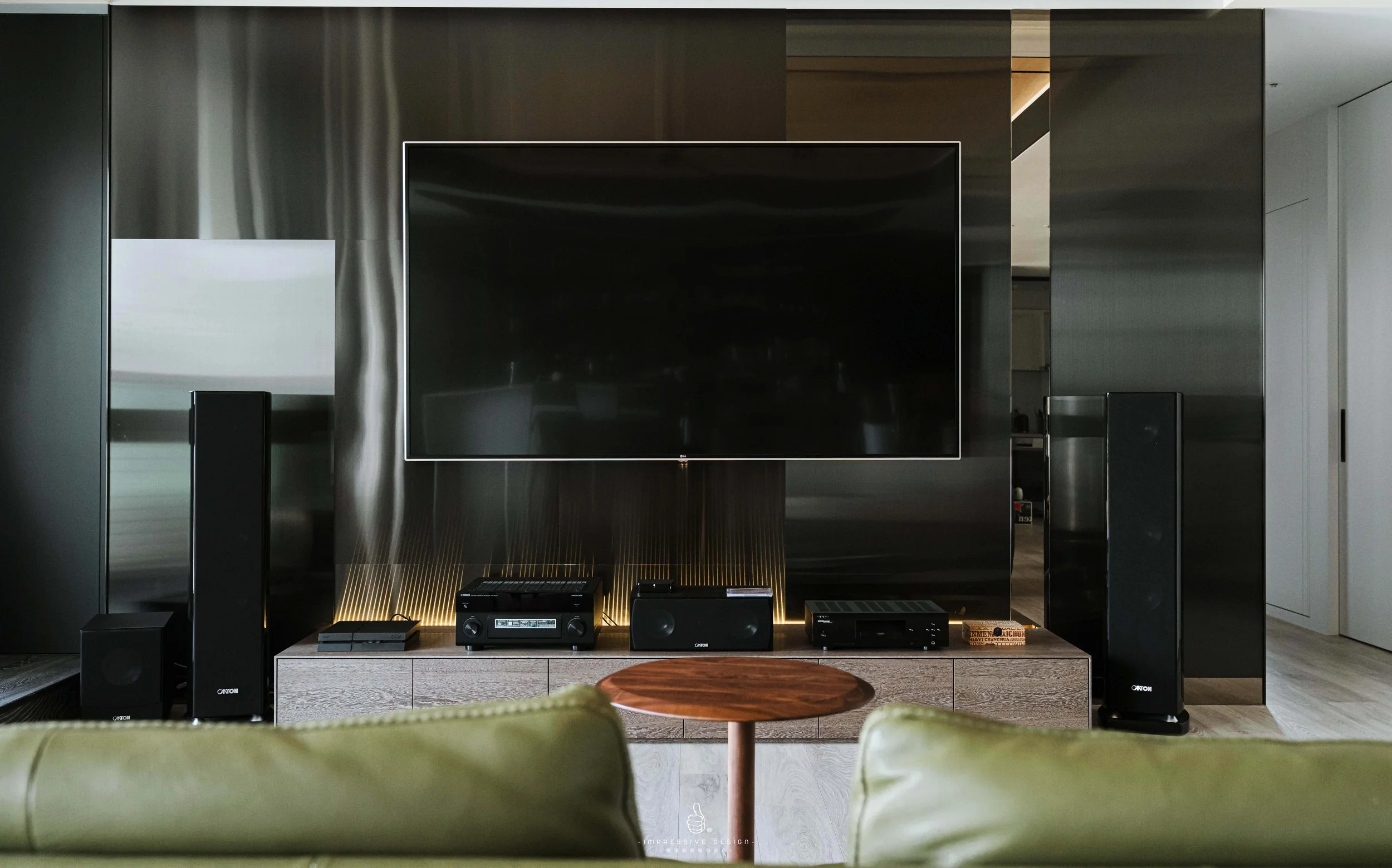 Modern living room with a large flat-screen TV, surround sound speakers, and media equipment on a wooden media console, with green couch in foreground.