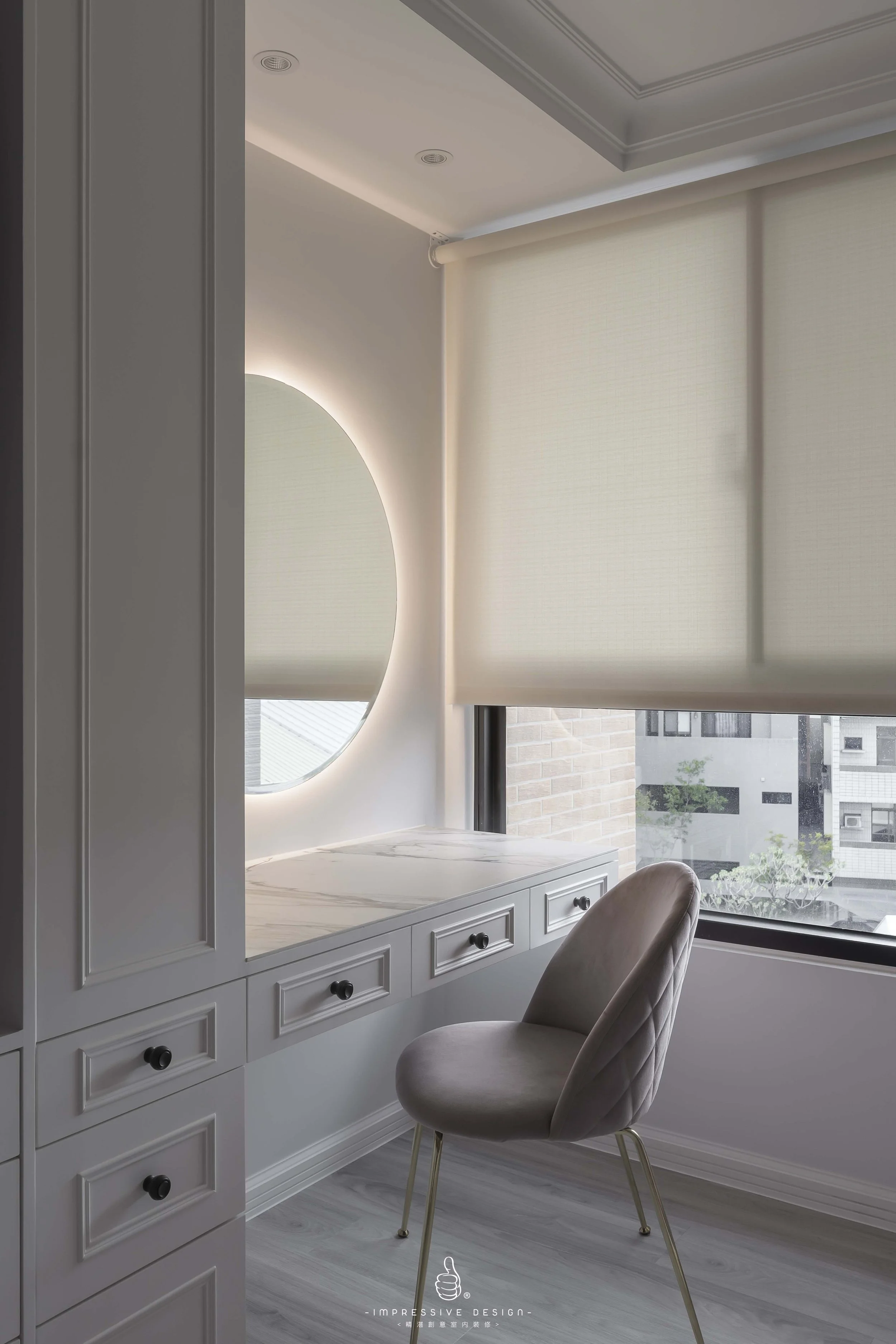 A light gray vanity desk with a marble top, positioned next to a large window with white blinds, a beige upholstered chair with gold legs, and a circular vanity mirror with backlighting, in a bright and modern room.