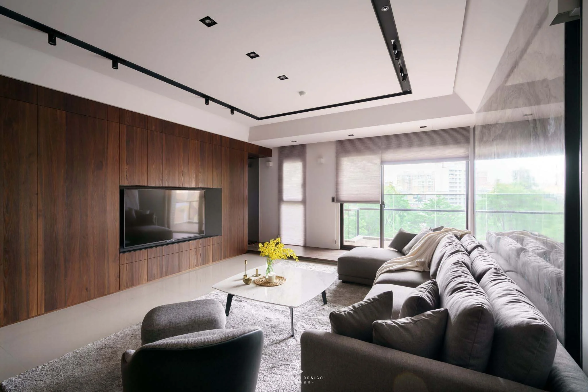Modern living room with a gray sectional sofa, a white coffee table with yellow flowers, a large window with blinds, and a wooden accent wall with a mounted flat-screen TV.