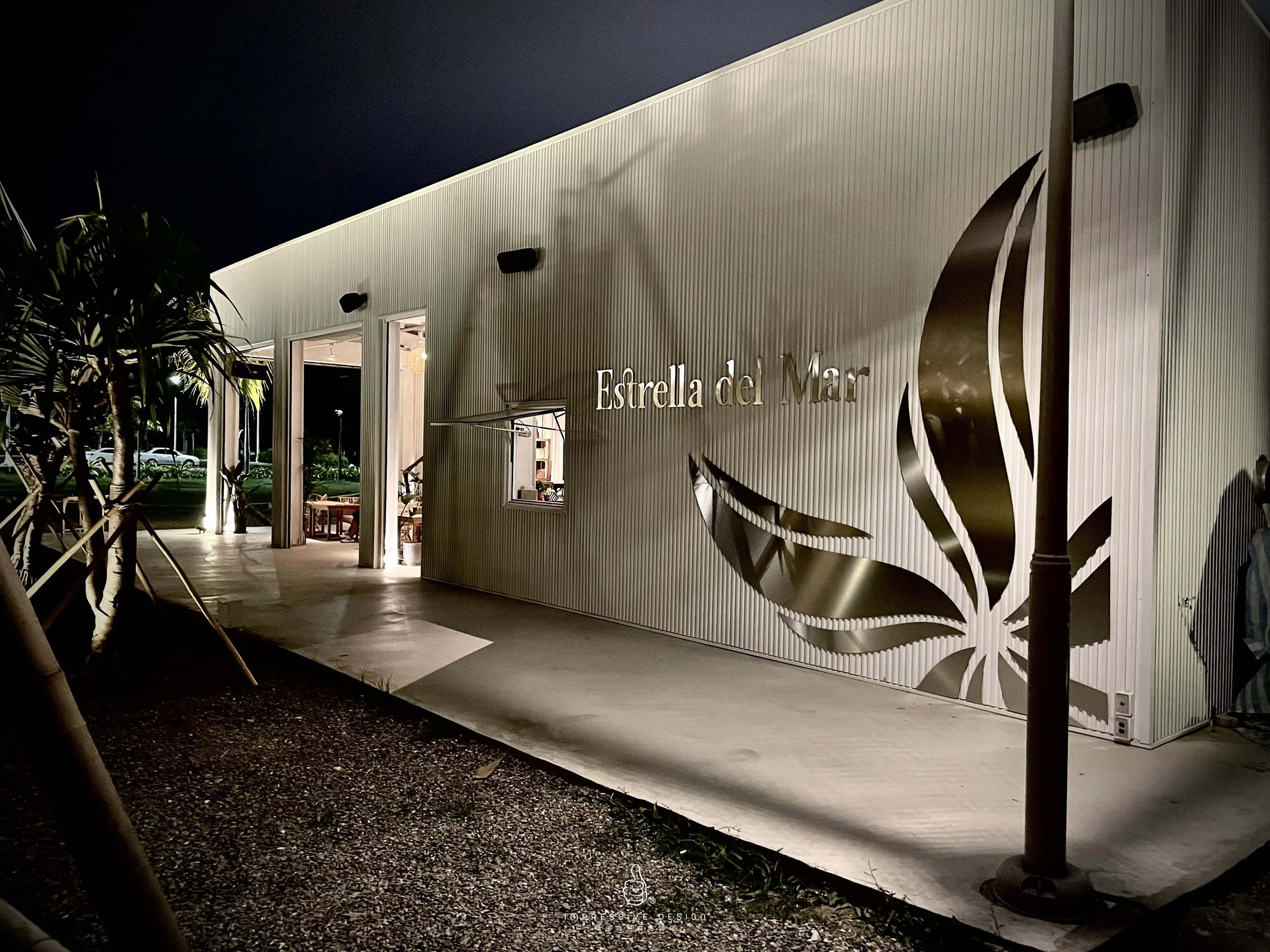 Night view of the exterior of a modern building with a metallic wall featuring a cutout design of a seashell and the text "Estrella del Mar" illuminated, with trees and parked cars visible in the background.