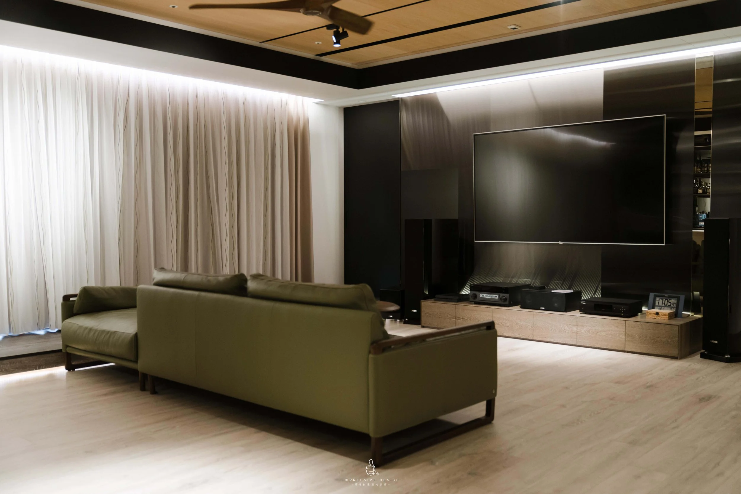 Modern living room with black accent wall, large flat-screen TV, olive green sofa, light wood flooring, white curtains, and built-in audio equipment.