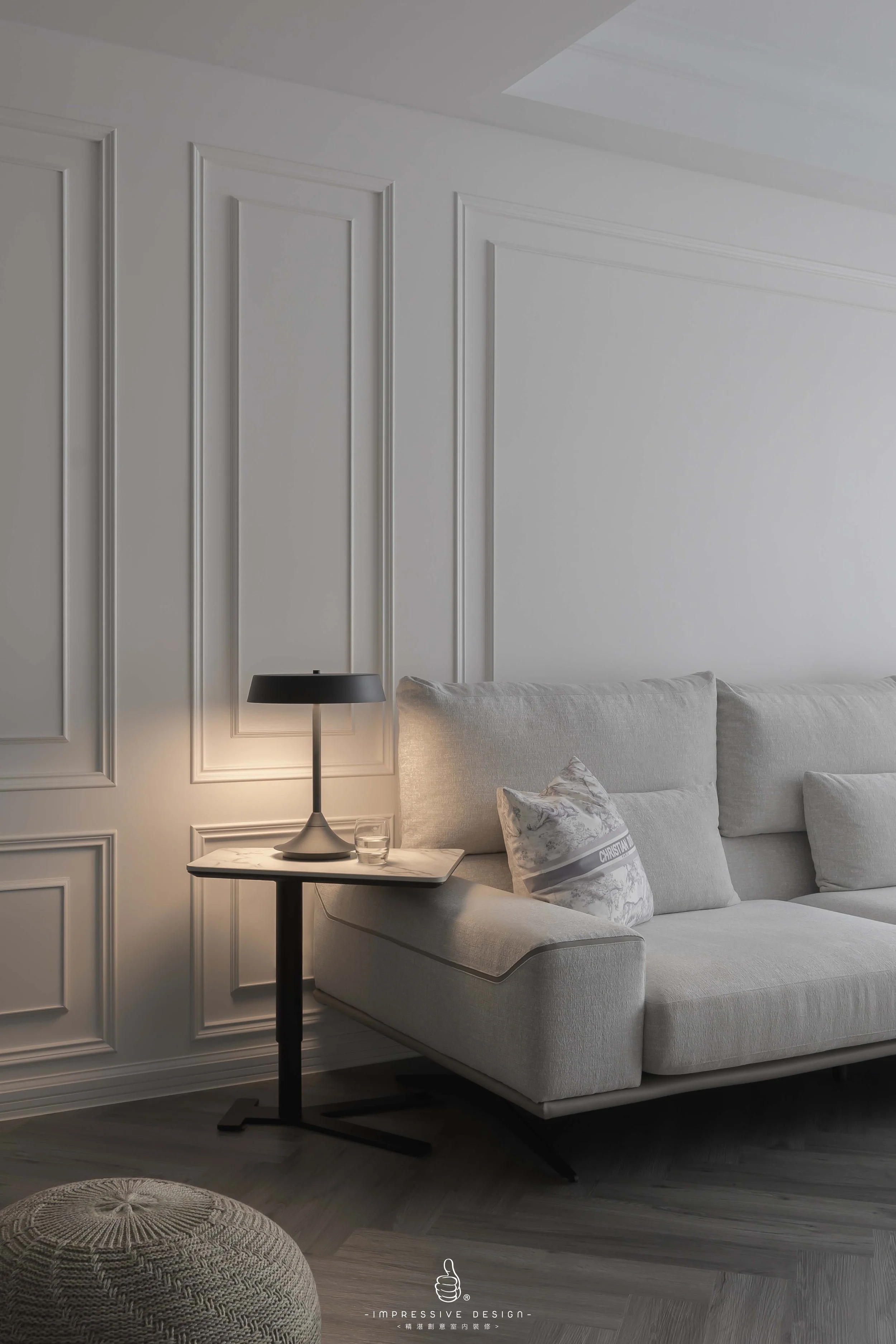A modern, minimalist living room corner with a light gray sofa, decorative cushions, a small side table with a black lamp, a glass of water, and a knotted pouf on a wood floor, against a white wall with decorative molding.