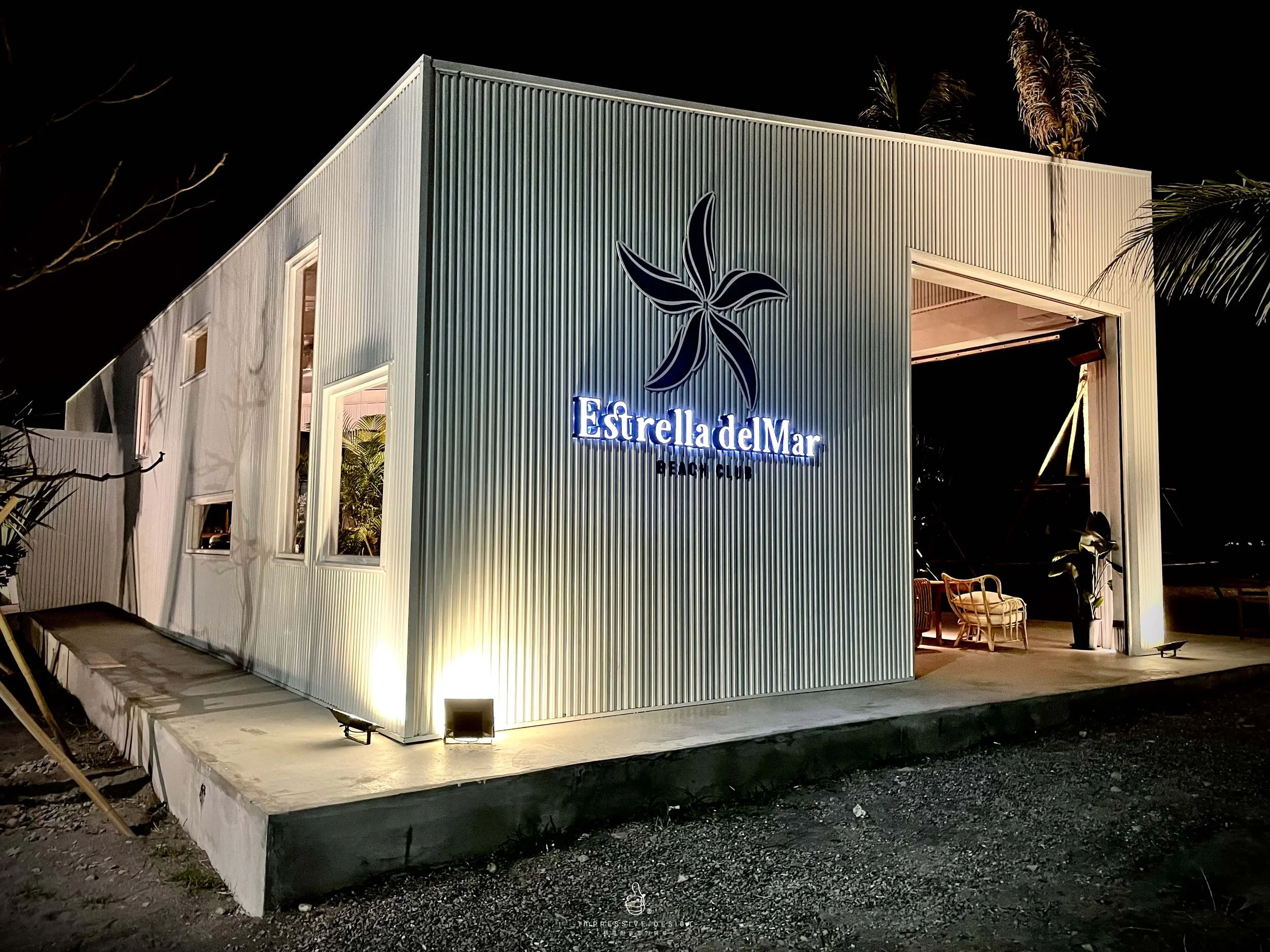 Night view of a modern building with a sign 'Estrella del Mar Beach Club' illuminated, featuring a large flower logo on the side, palm trees, and outdoor seating visible inside.