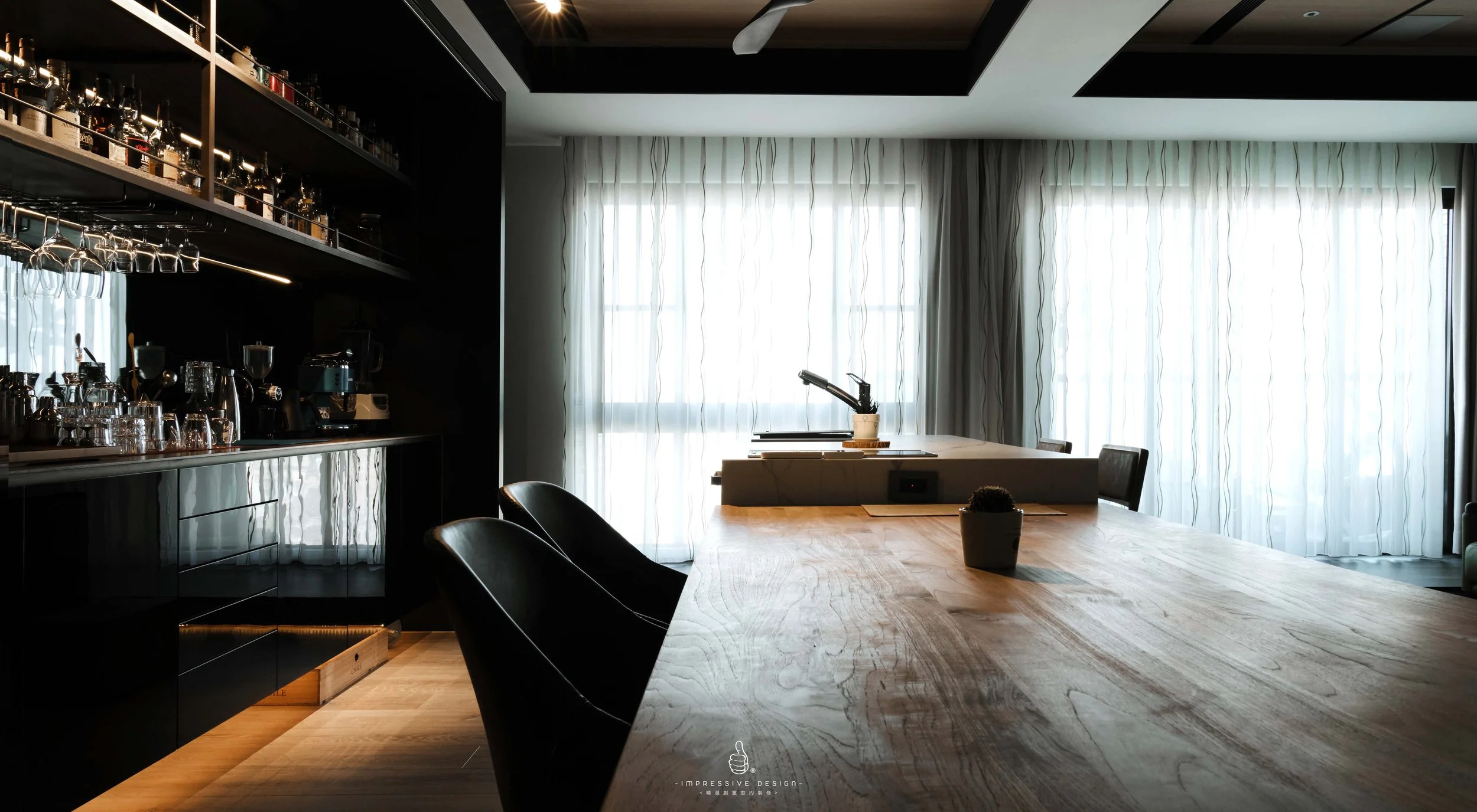 Modern kitchen with a large wooden dining table, black chairs, a window with sheer curtains, built-in black cabinetry, and a countertop with kitchen appliances.
