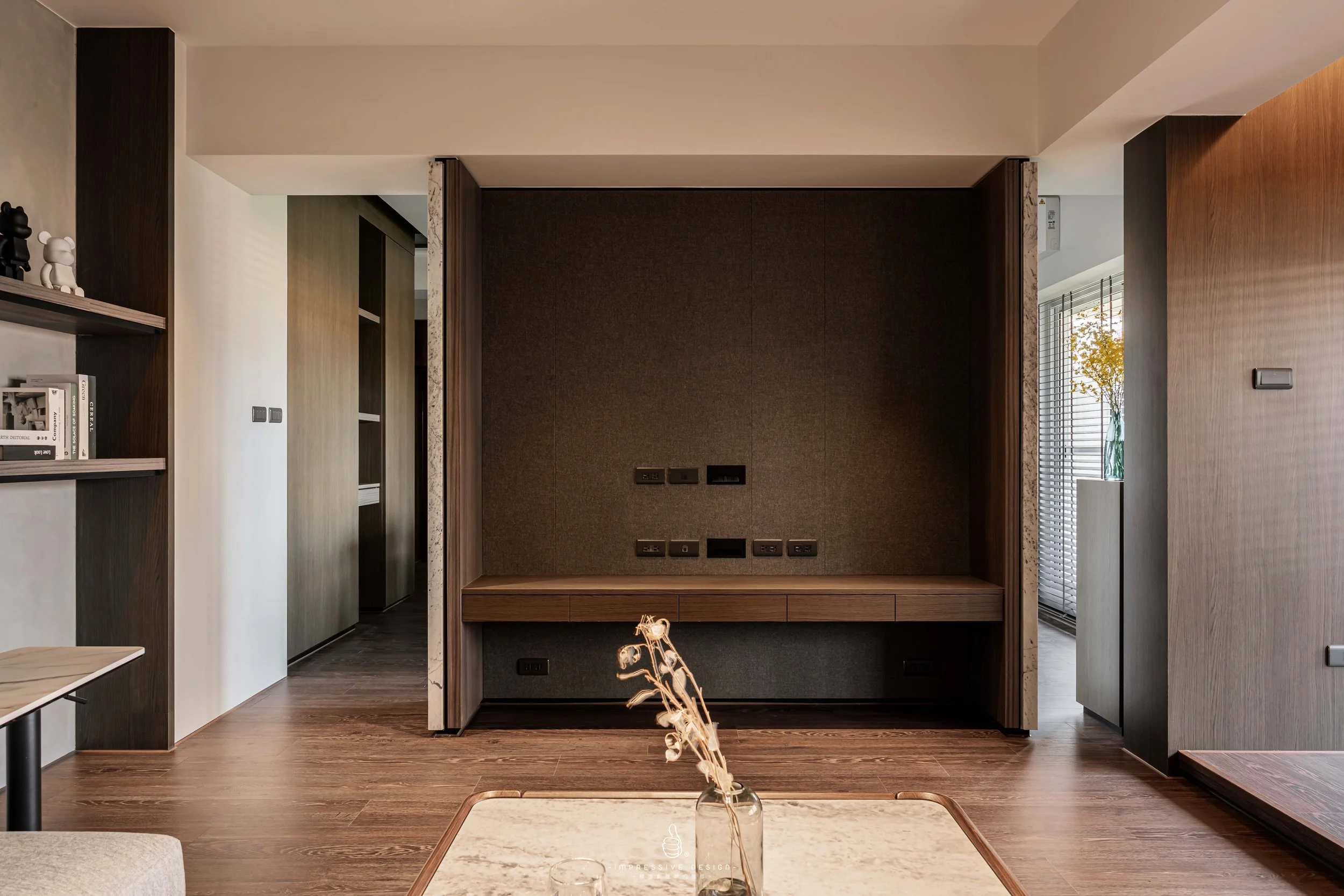 Modern living room with wooden flooring, built-in shelves, and a wall with multiple electrical outlets and ports, with a vase and dried plant on a table in foreground.