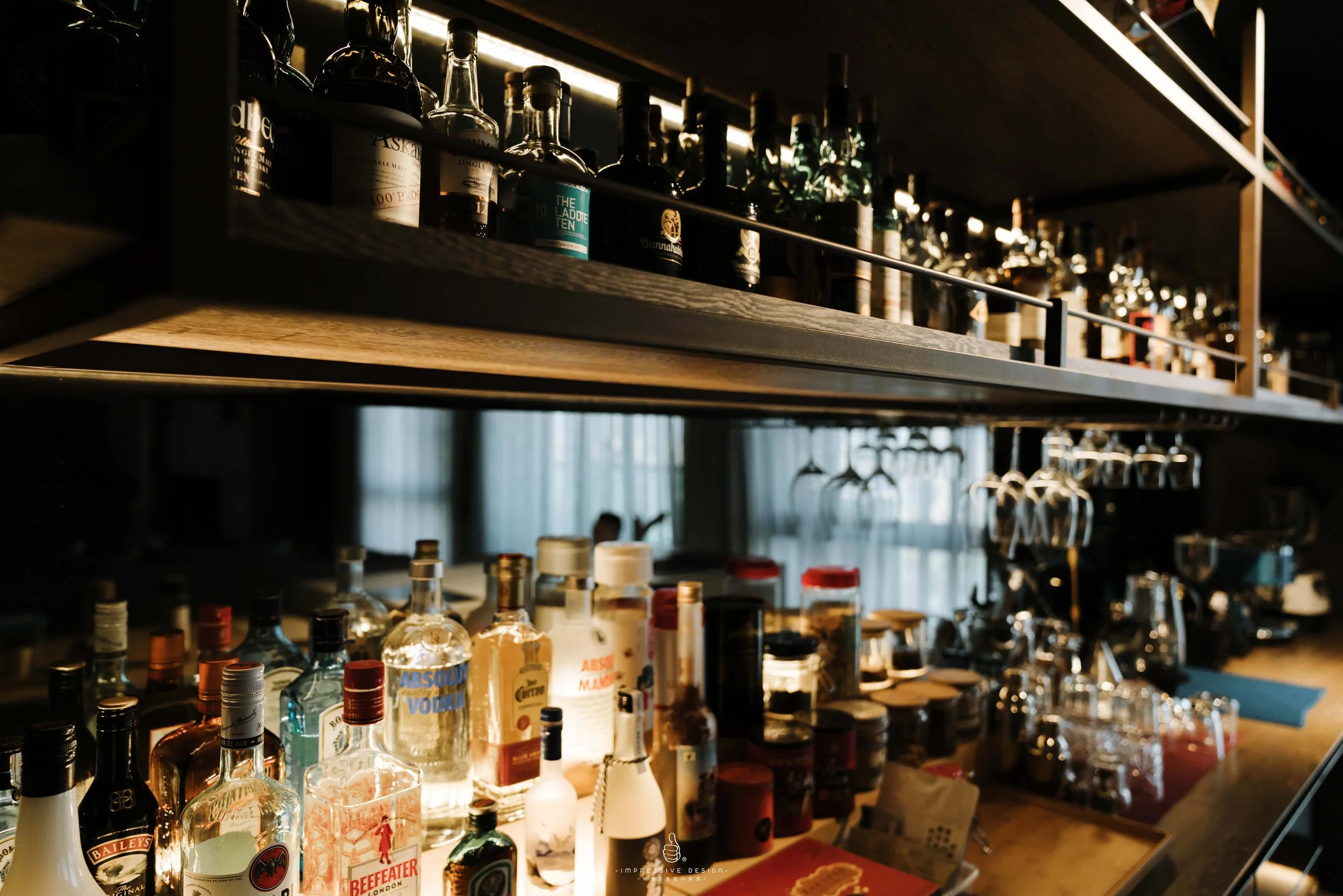 A bar shelf stocked with liquor bottles, glasses, and bar tools illuminated by warm lighting.