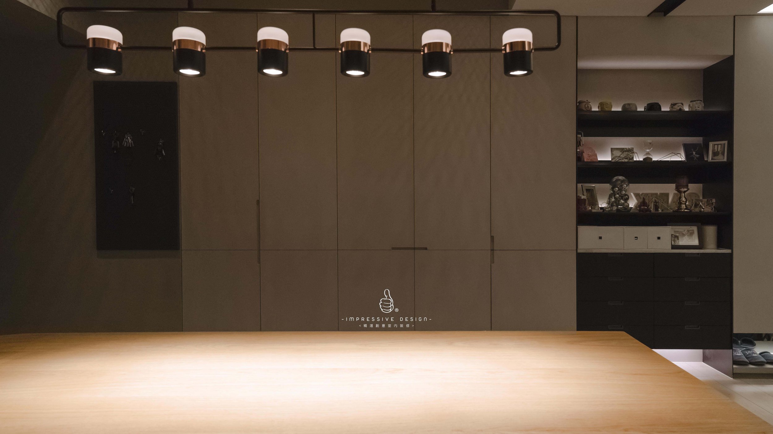 A modern interior with a wooden table in the foreground, a beige cabinet wall, a black shelving unit with decorative items, and modern ceiling lights.