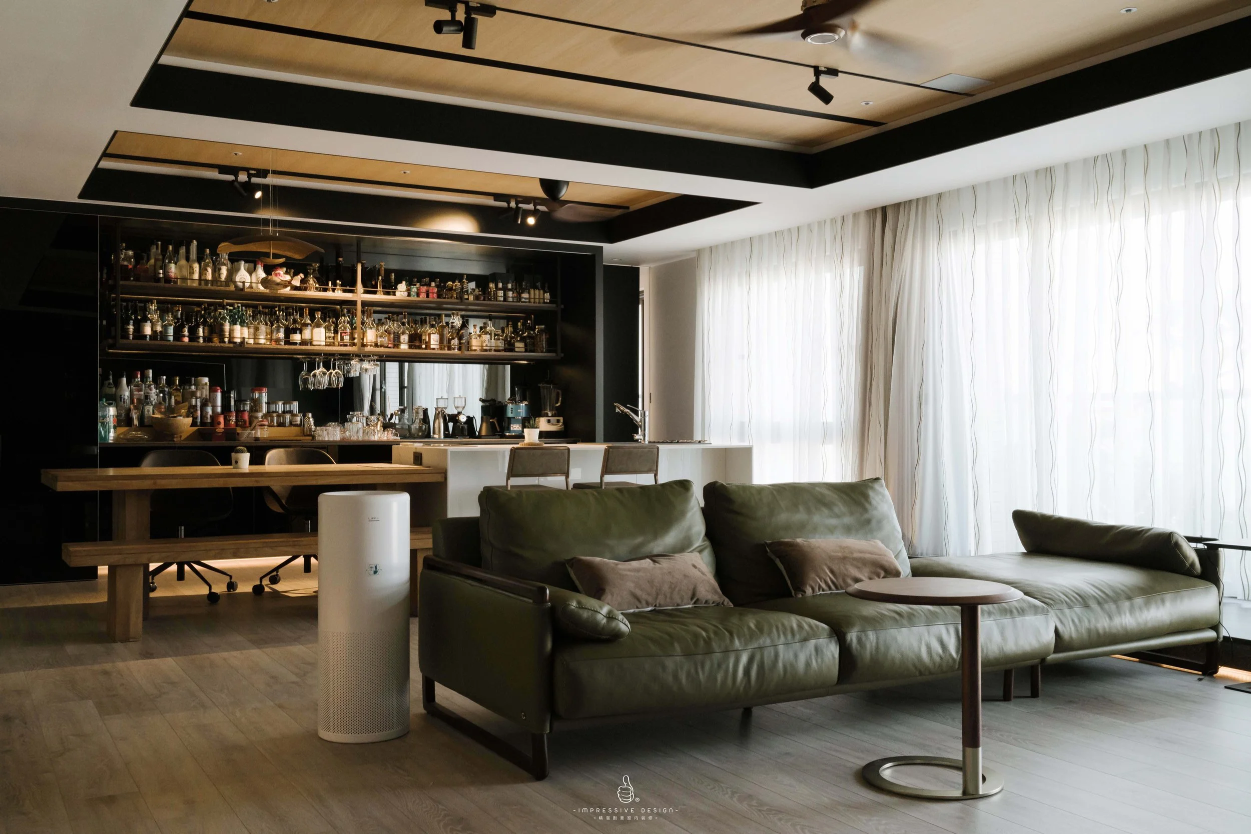 Modern living room with a green leather sofa, a small round wooden side table, large windows with white curtains, a bar area with bottles on shelving, and chairs in front of the bar, in a well-lit space.