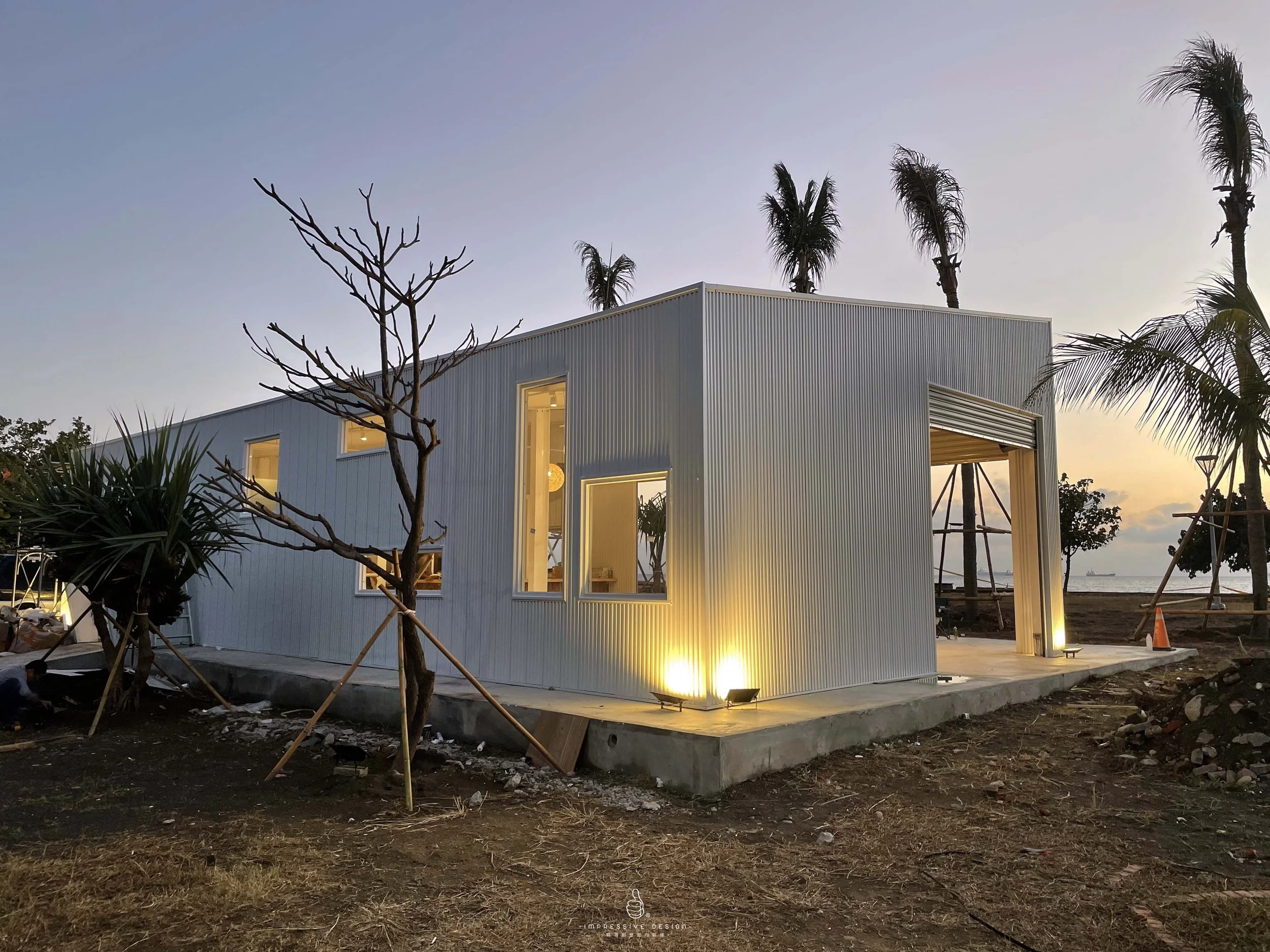 Modern house with corrugated metal exterior walls, illuminated from the outside, situated in a tropical landscape at dusk with palm trees and a view of the ocean in the background.
